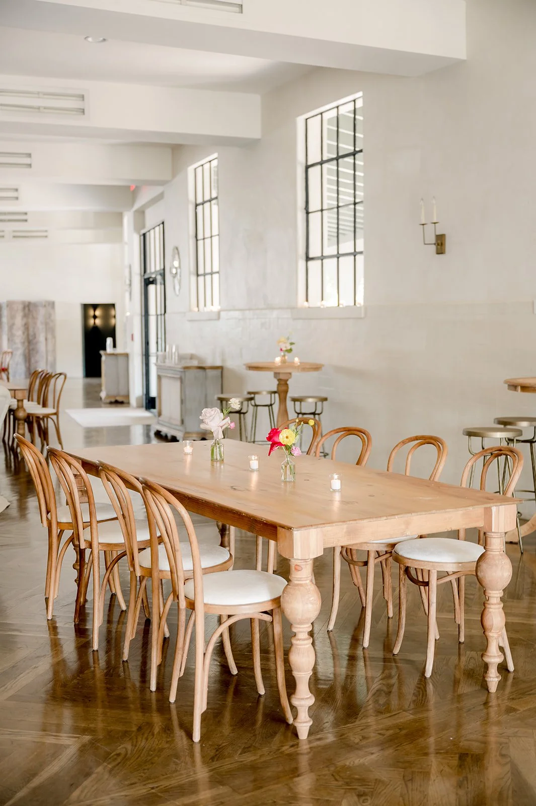 A bright, minimalist dining area with a large wooden table, surrounded by wooden chairs with white cushions, adorned with small vases of flowers and lit candles, featuring high windows and white walls.