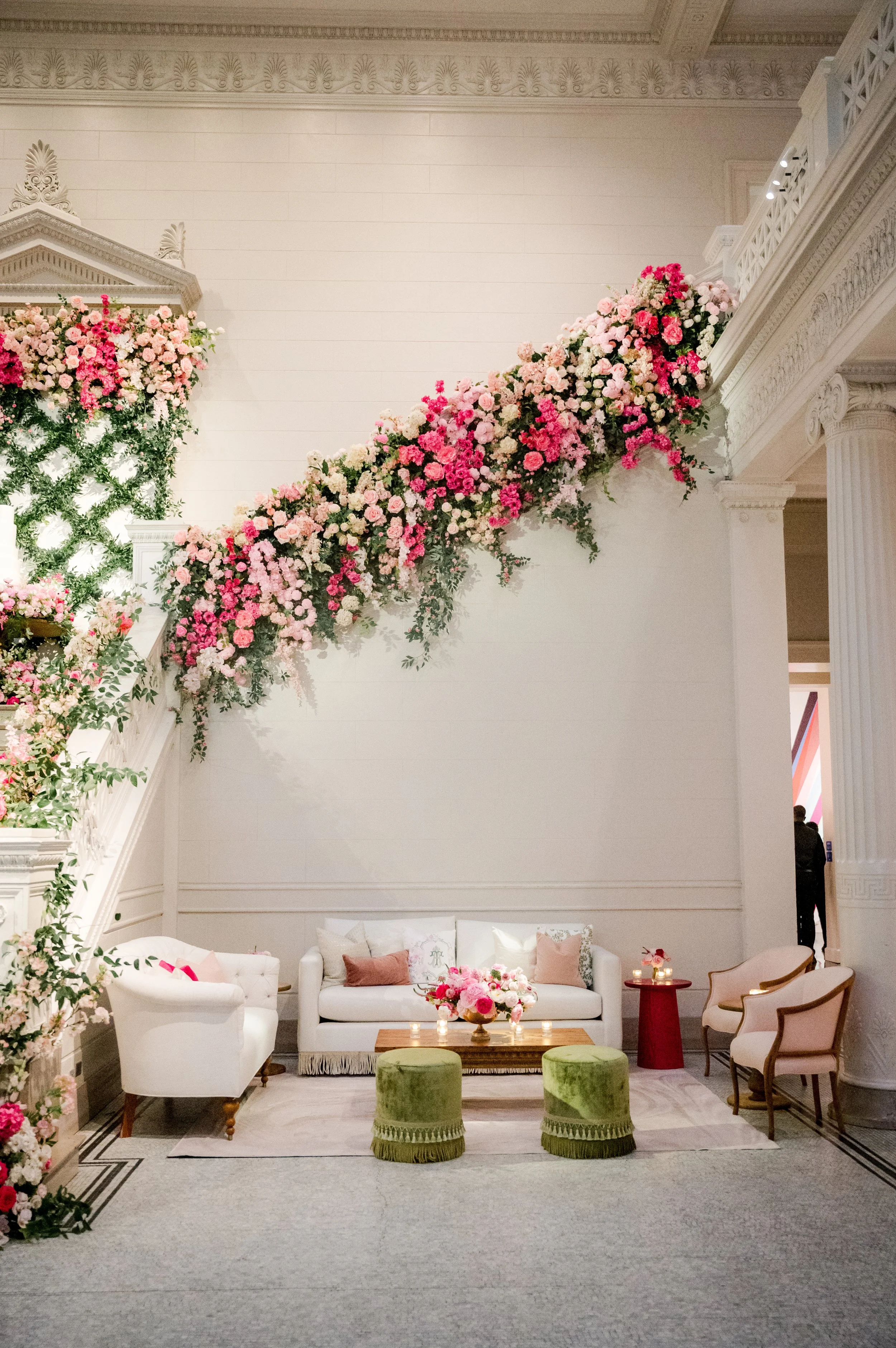Elegant lounge area decorated for a wedding with a floral arrangement cascading from the upper level to the main floor, pastel-colored cushions on white sofas, a wooden coffee table with pink flowers, and two green velvet stools in front. The backgro