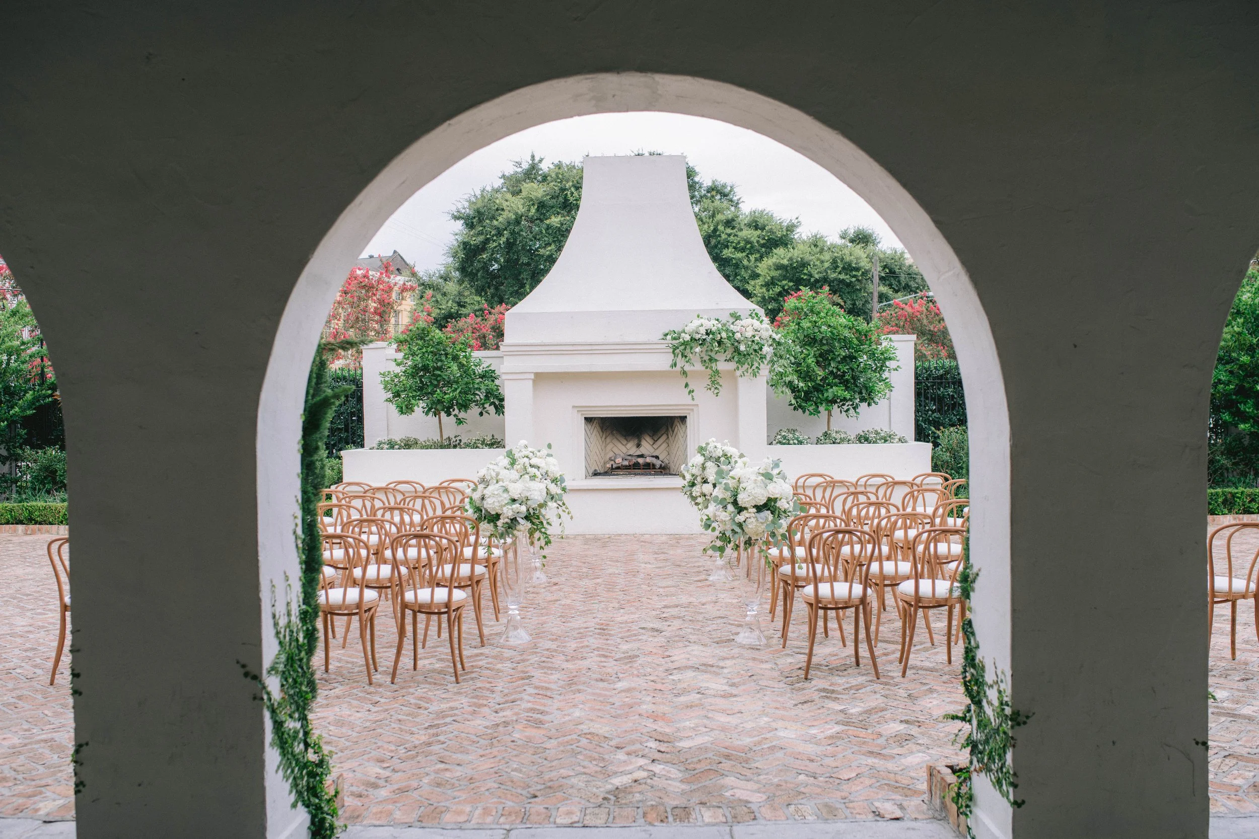 Outdoor wedding ceremony setup with chairs and floral arrangements seen through an arched opening, with a white fireplace-like structure in the background surrounded by greenery.
