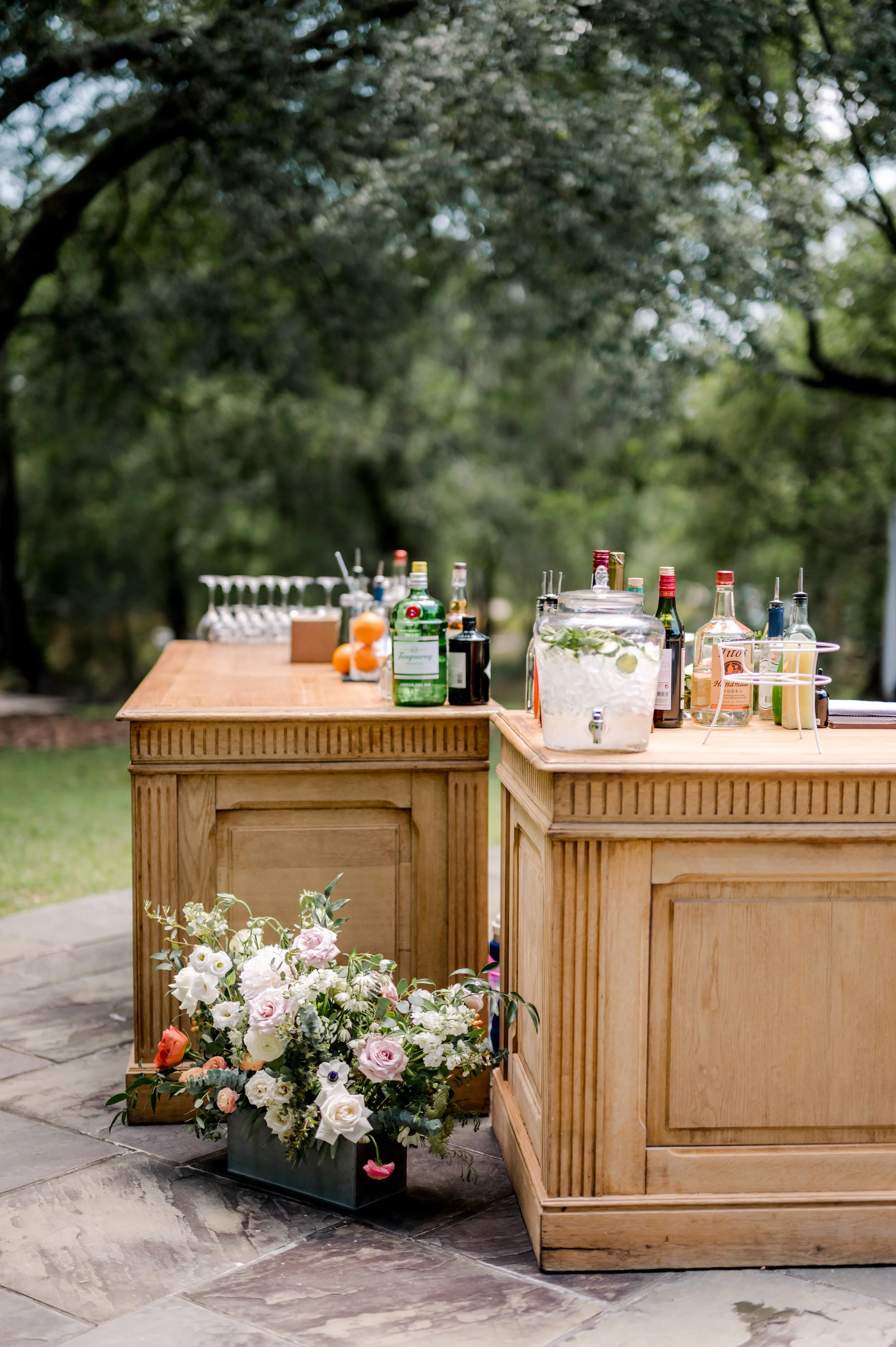Two wooden tables set up outdoors with bottles of alcohol, mixers, and glassware for a celebration, with a large bouquet of flowers in the foreground and trees in the background.