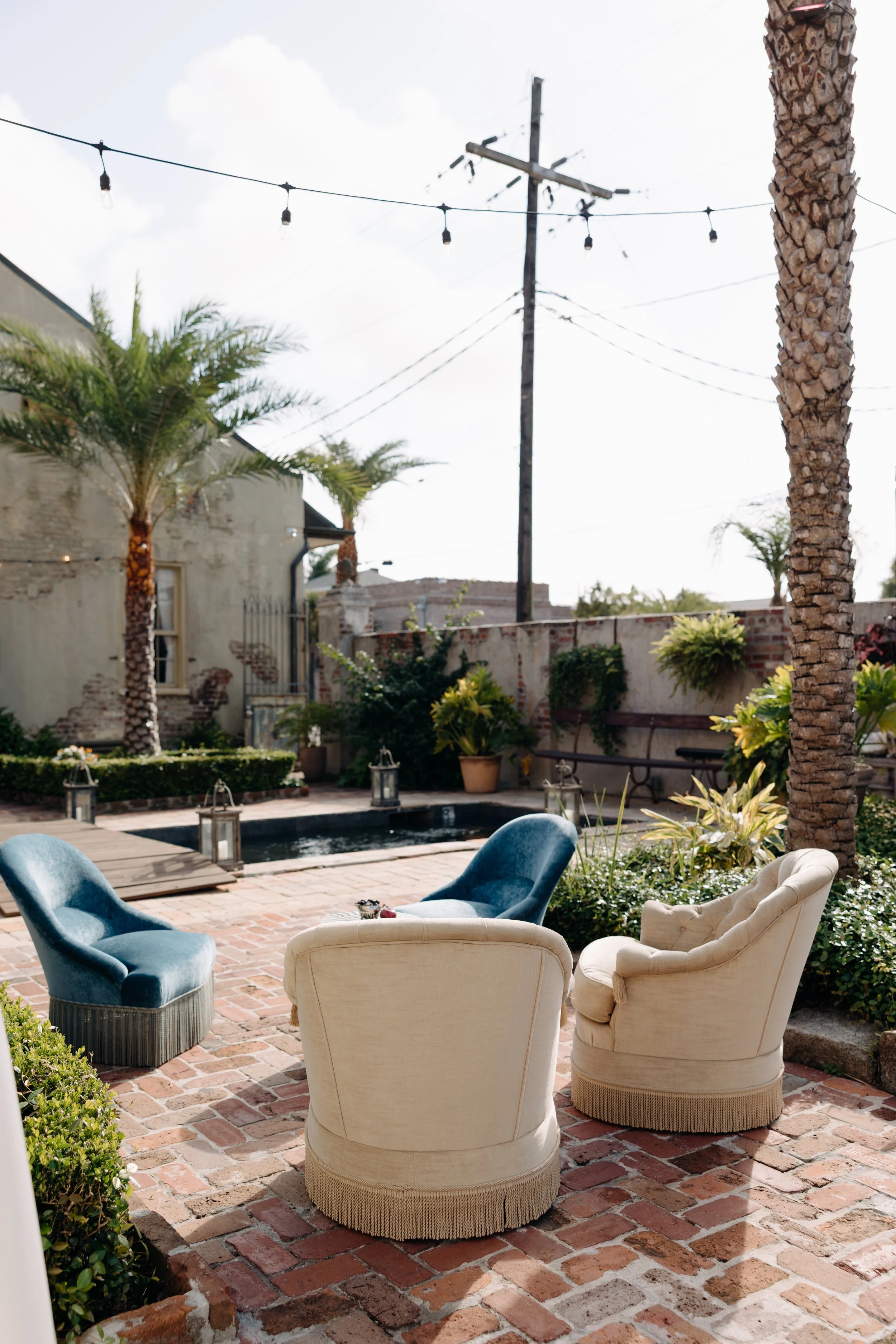 An outdoor patio area with vintage white and blue armchairs on a red brick floor, surrounding a small fire pit, with palm trees and potted plants nearby, a brick wall in the background, and string lights hung overhead.