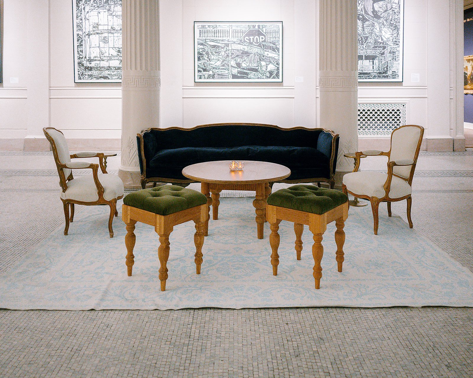 Elegant seating area with a black velvet sofa, two white upholstered armchairs, two green cushioned ottomans, and a round marble-top table with candles, set on a light blue patterned rug in a gallery or museum with white walls and framed black and wh