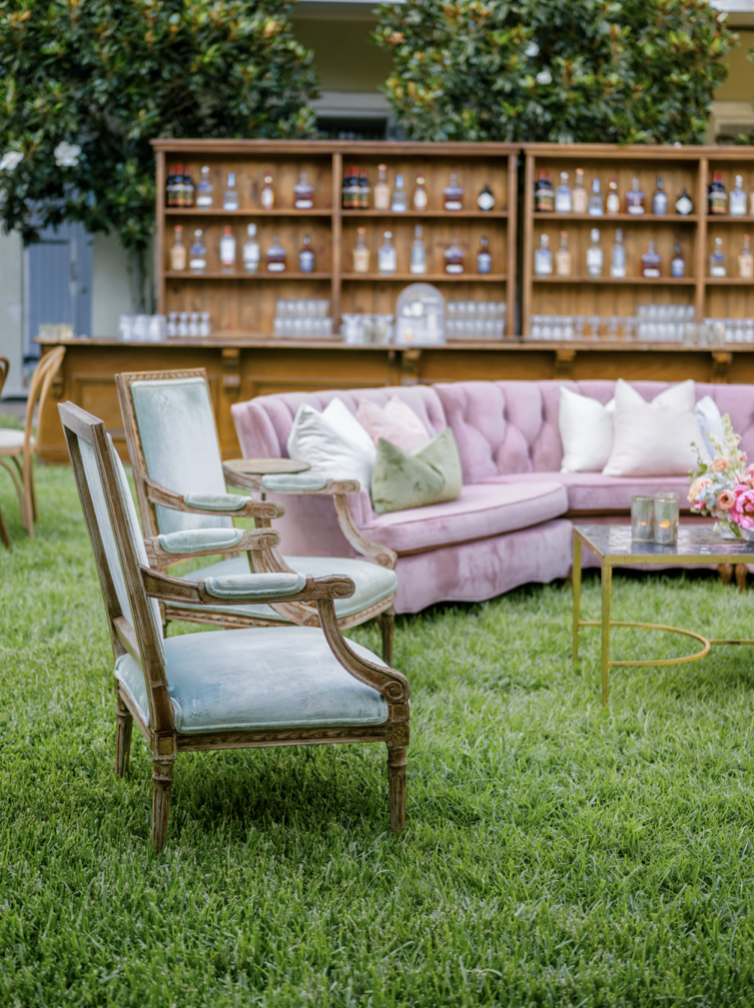 Outdoor seating arrangement with vintage armchairs and a pink velvet sofa on a grassy lawn, with a wooden shelf of liquor bottles and trees in the background.
