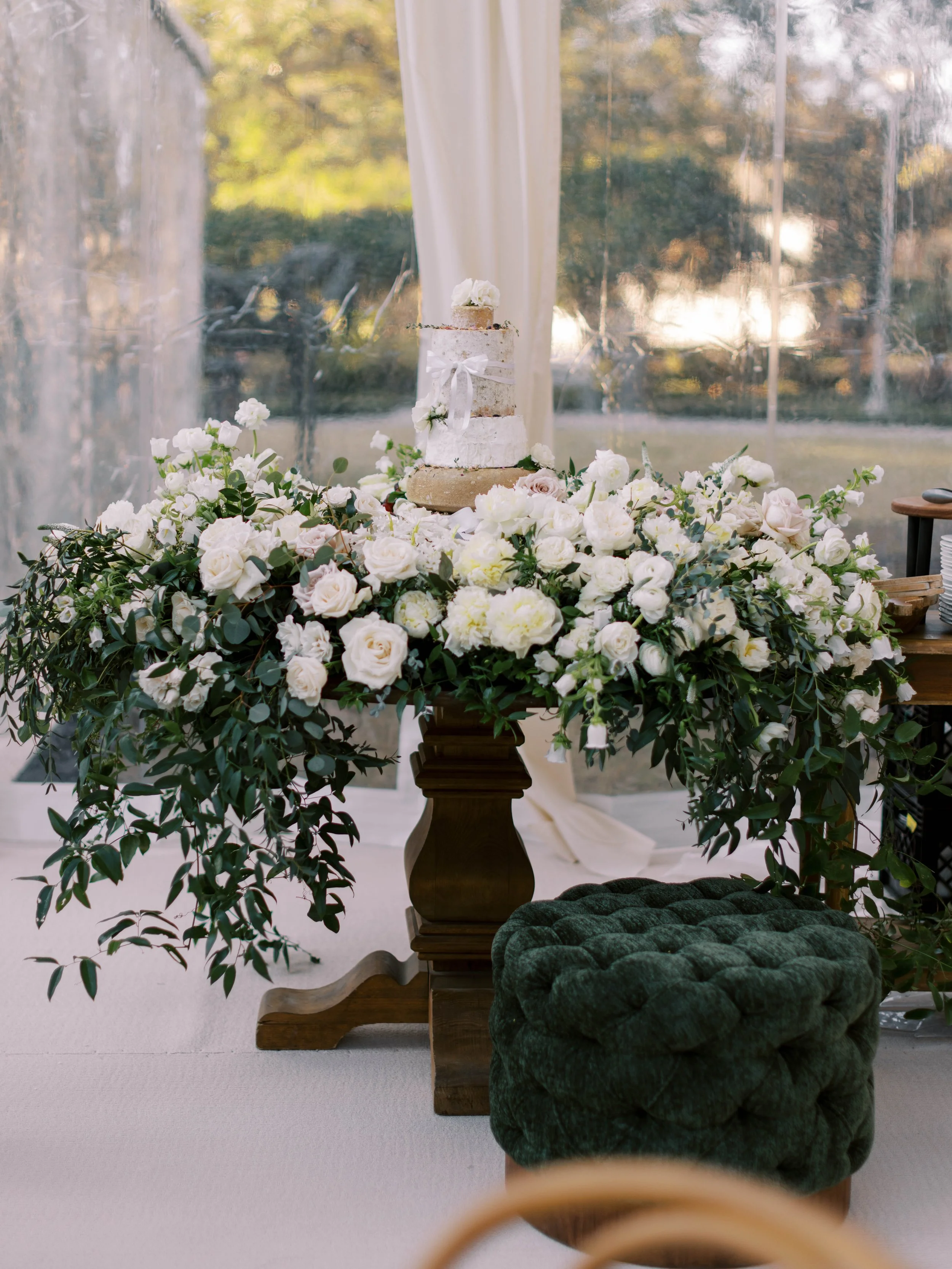 A wedding cake surrounded by white flowers and greenery on a wooden stand, with a sheer curtain and trees visible outside through a window in the background.