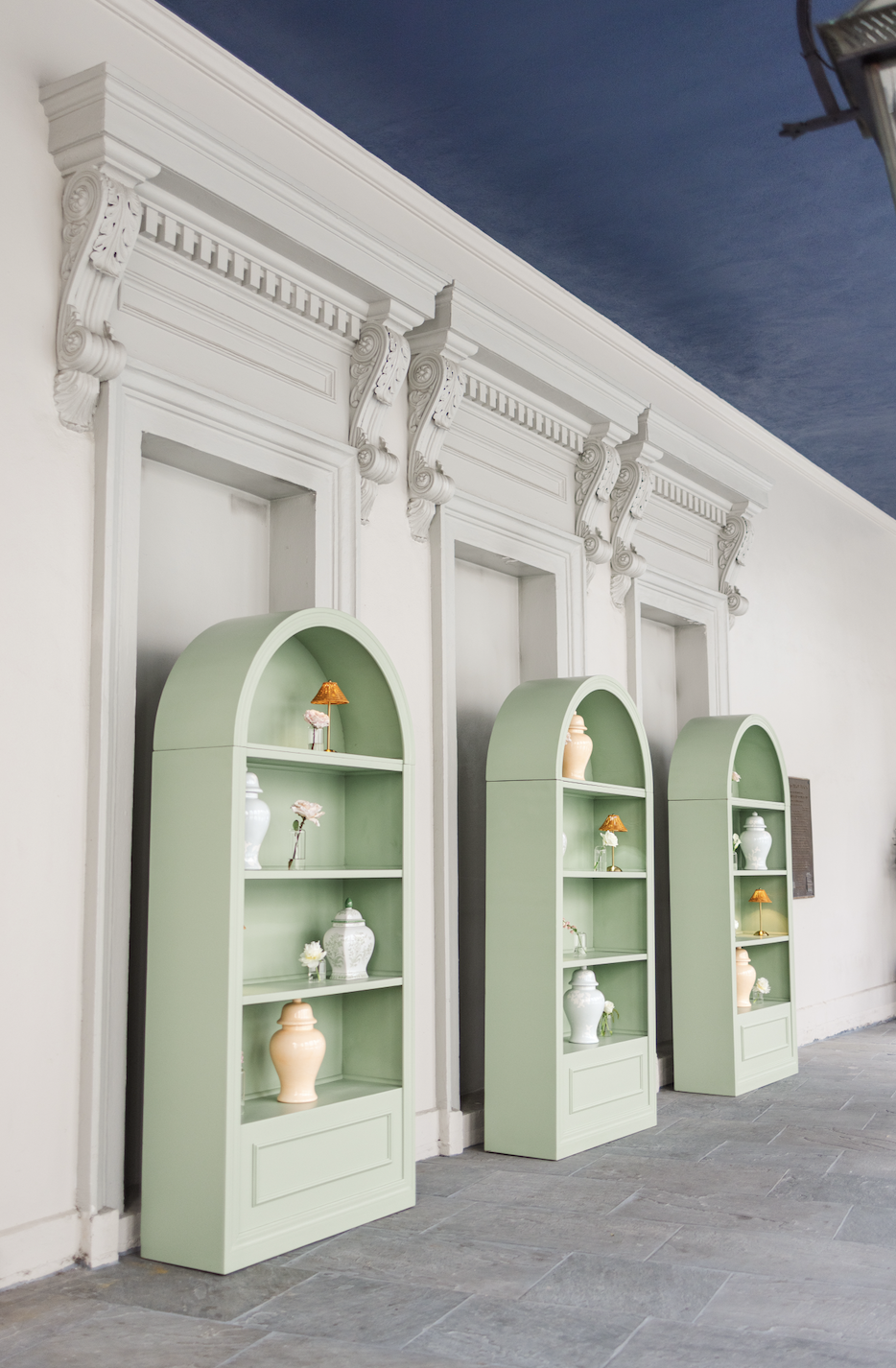 Three light green arched display shelves with decorative vases and flowers against a white wall with ornate molding, and a dark ceiling.