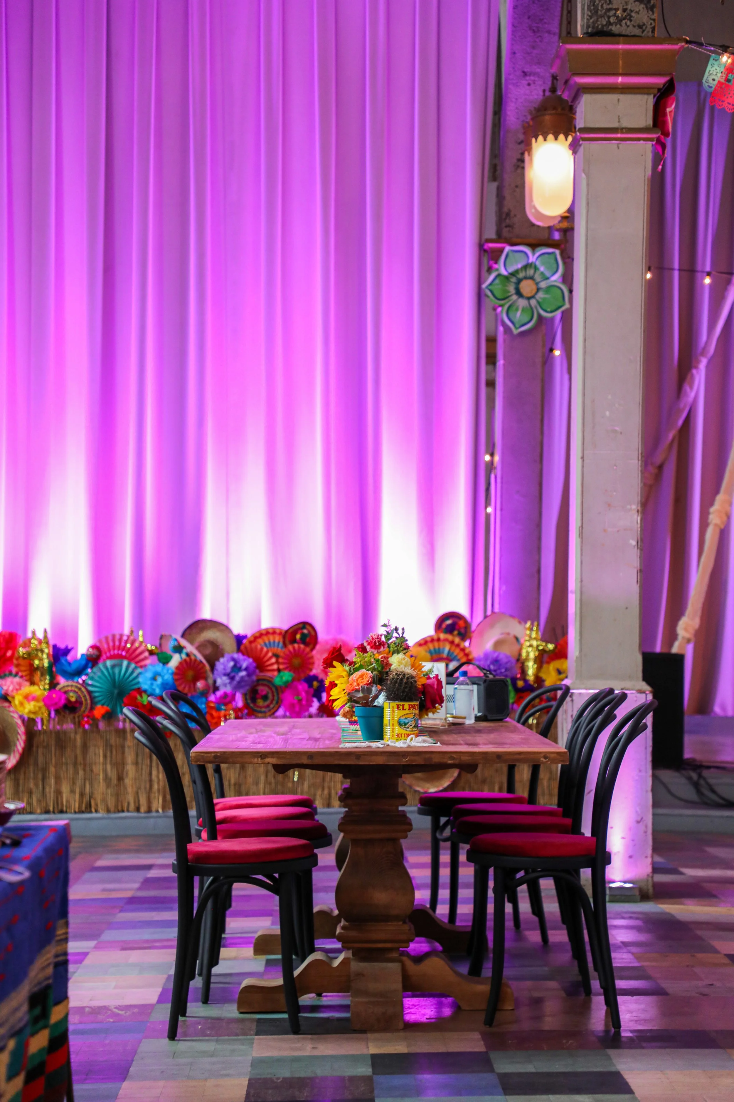 Indoor celebration setting with a wooden table surrounded by black chairs with red cushions, decorated with colorful flowers and party favors. The background features purple lighting, pink and purple curtains, and decorative floral and lantern accent