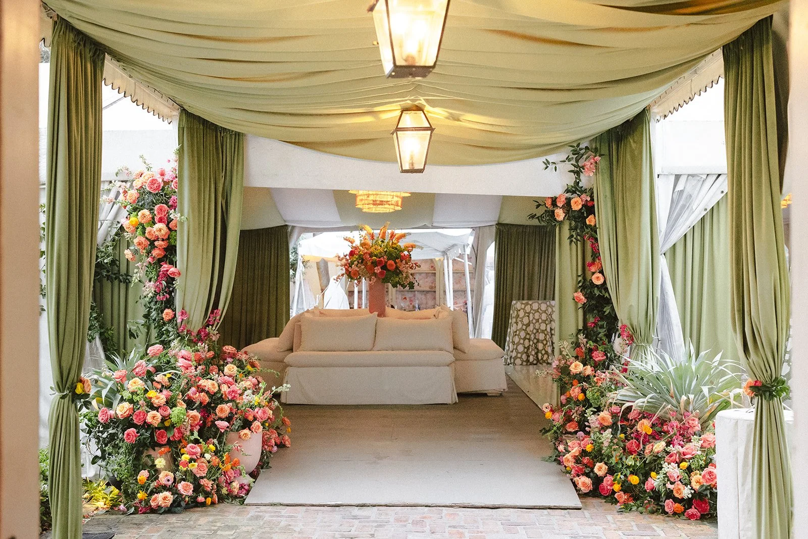 A decorated venue with green curtains, floral arrangements, and a central sofa with a large bouquet of flowers on it, set up for a wedding or celebration.