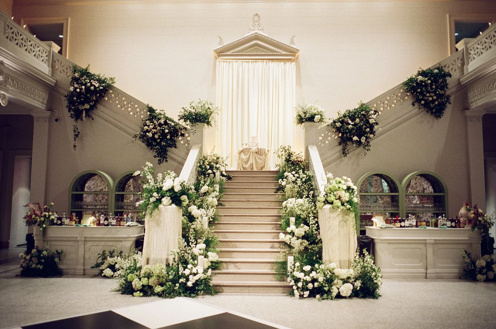 Elegant wedding altar with floral arrangements on stairs and at base, draped fabric backdrop, and decorative elements in a grand indoor setting.