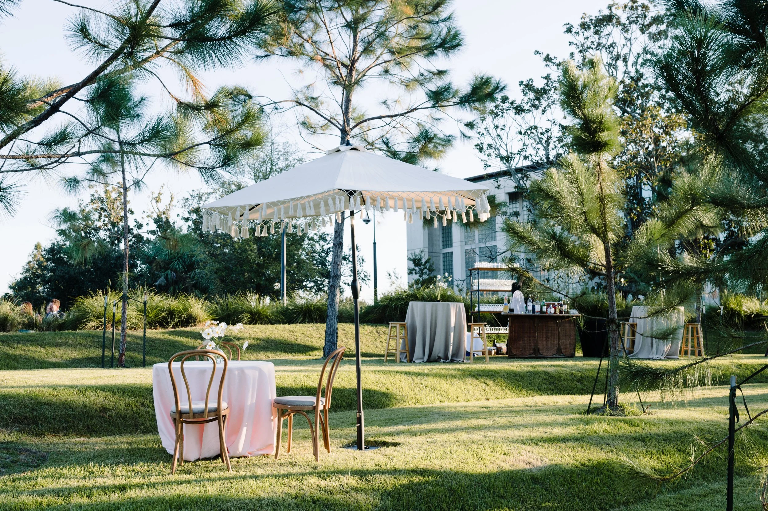 Outdoor event setup with round tables, chairs, a white patio umbrella, and a bar area, surrounded by trees and greenery.