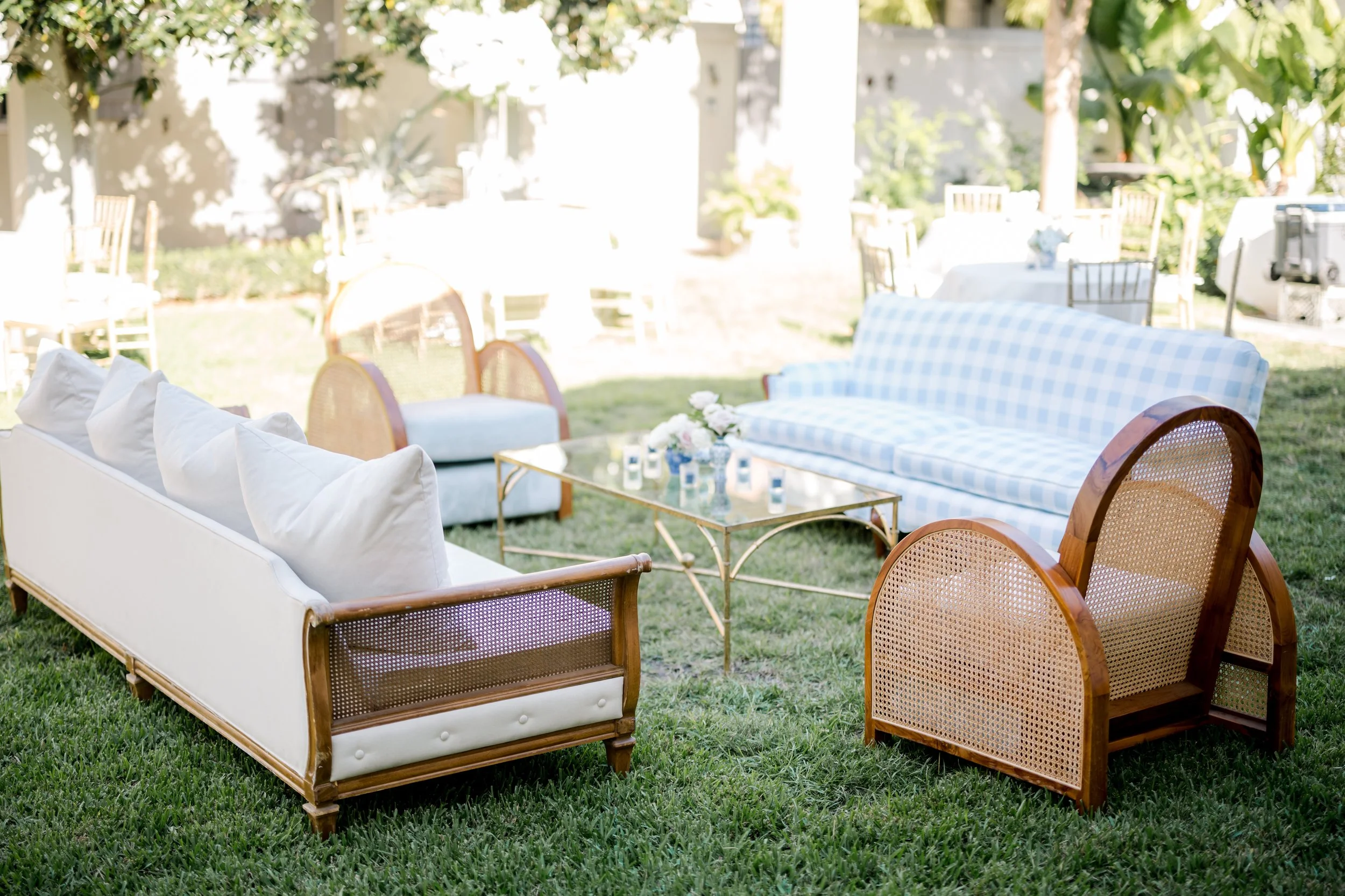 Outdoor patio setup with white and patterned sofas, wooden chairs, glass coffee table, and dining tables with chairs, surrounded by green trees and grass.