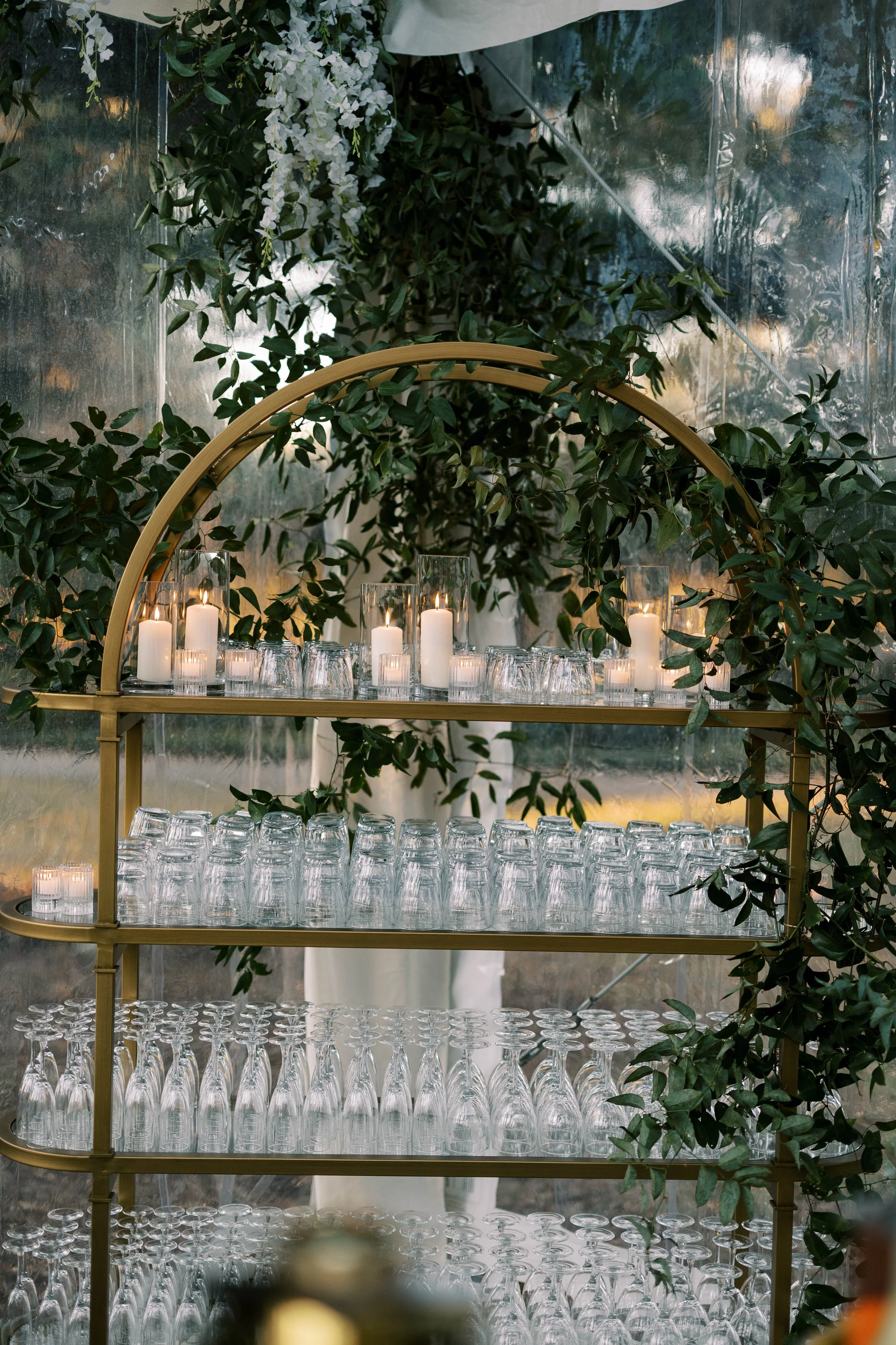 A multi-tiered metal bar cart with glassware and candles, decorated with green leaves and white flowers in a greenhouse or similar setting.