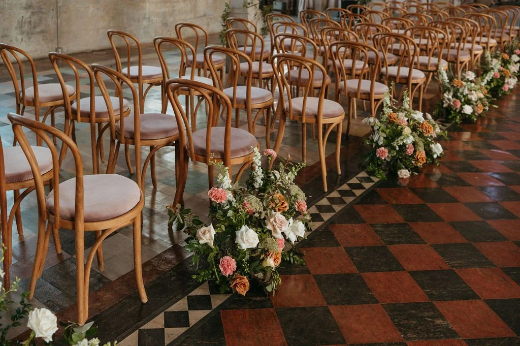 Wedding ceremony setup with rows of wooden chairs and floral arrangements along the aisle on a checkered wooden floor.
