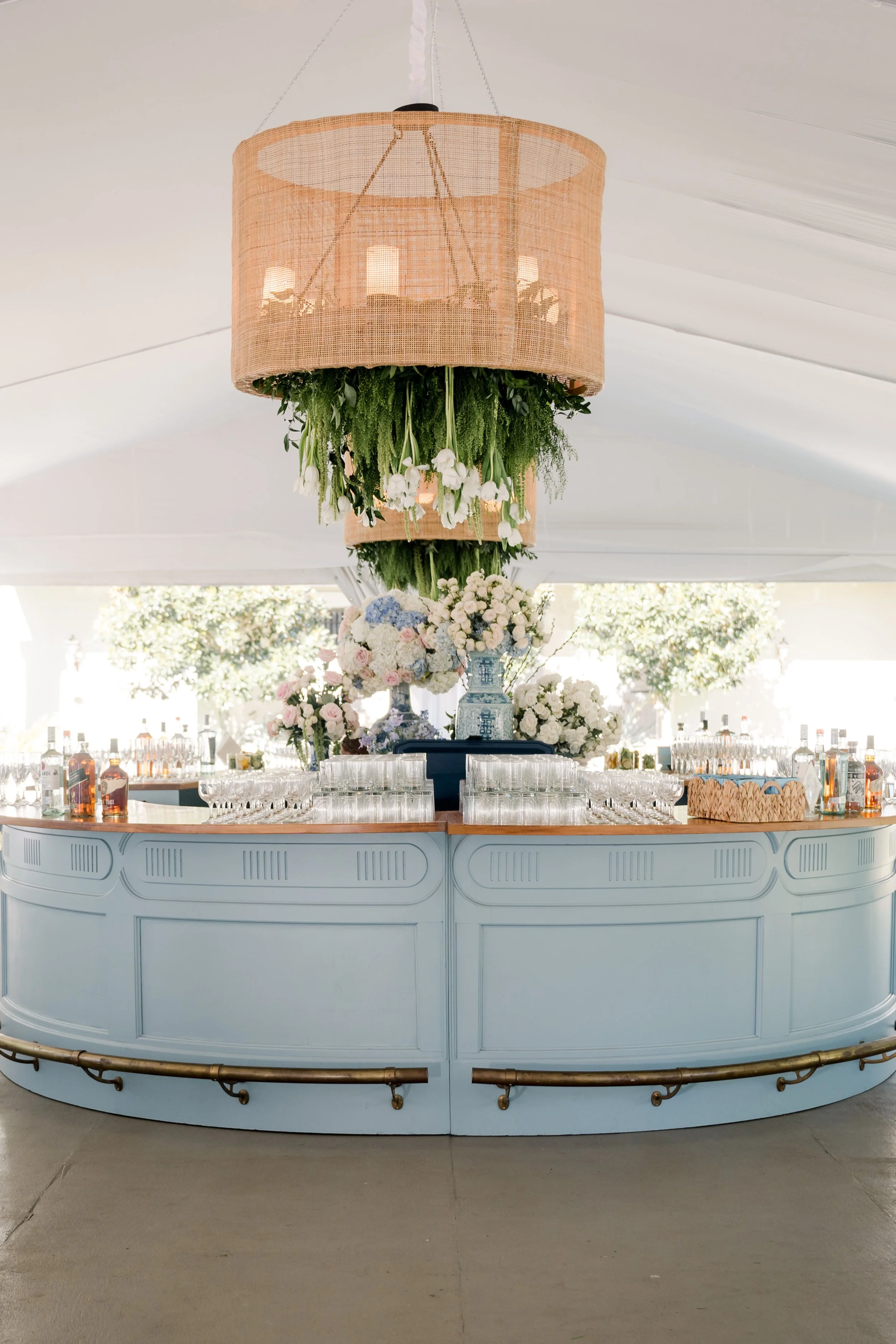 A bar with a large floral arrangement on top, decorated with greenery and white flowers. Above the bar hangs a woven, circular pendant light with plants inside. The background shows a white tent ceiling and outdoor trees.