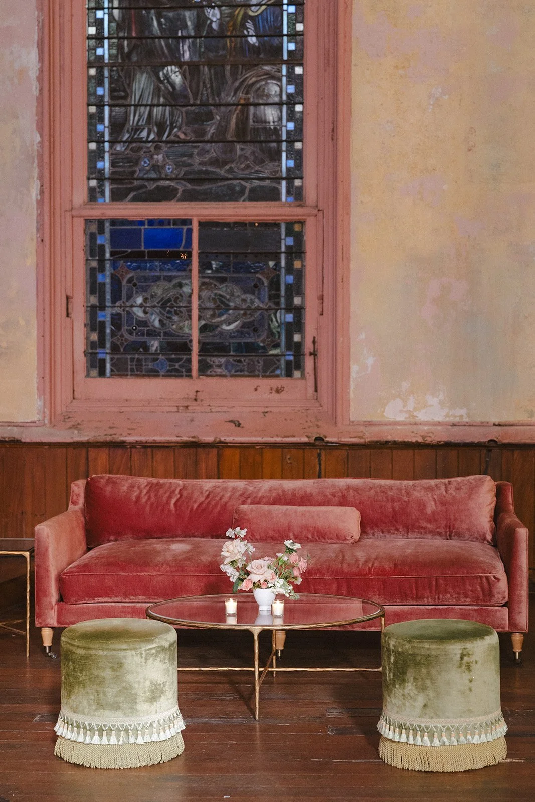 A vintage room with a pink velvet sofa, green velvet footstools with decorative fringes, a small glass top coffee table, a flower arrangement, a candle, a tall stained glass window, and peeling yellow walls.