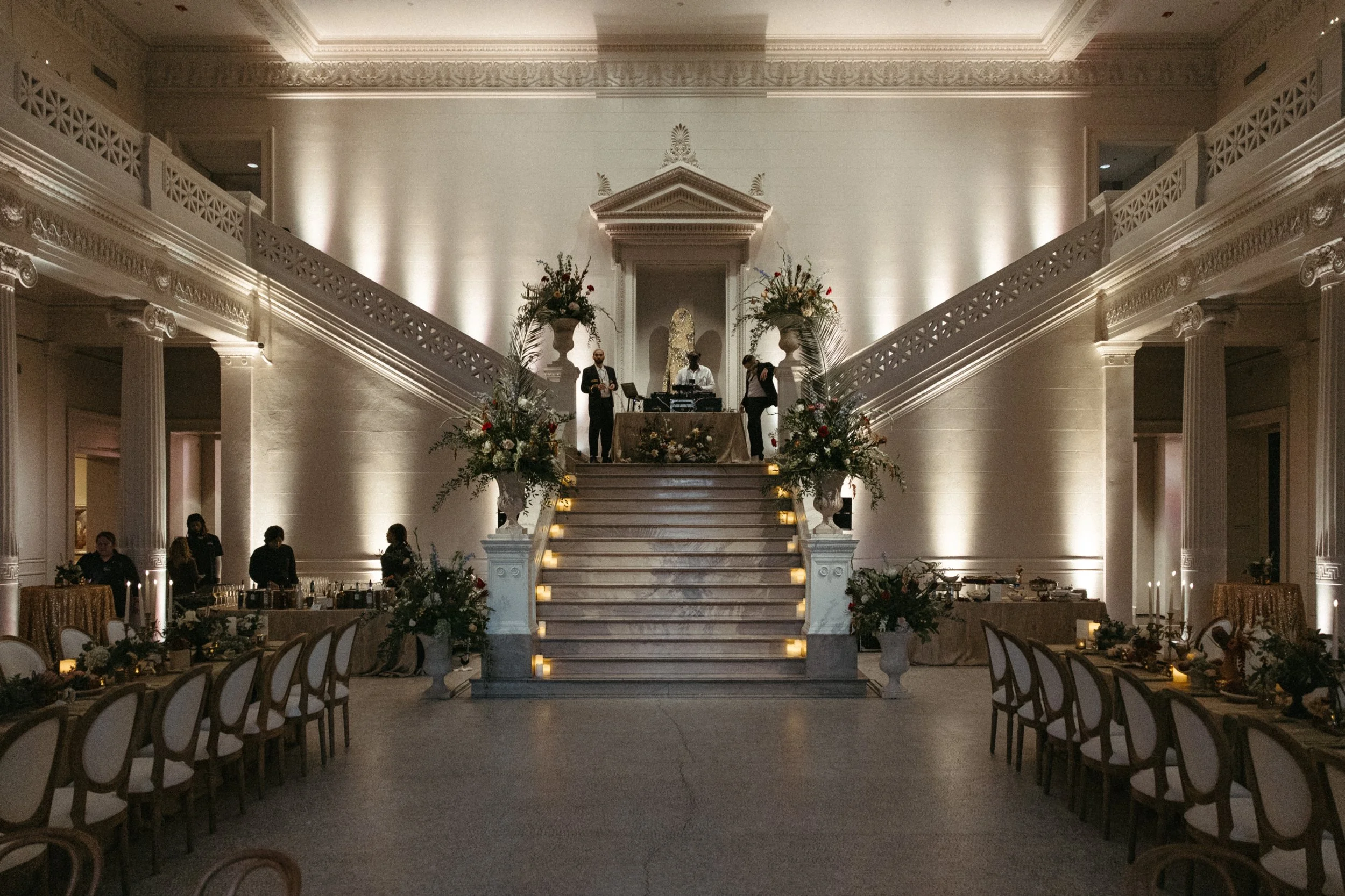 Luxury event hall with grand staircase, floral arrangements, and a DJ setup at the top of the stairs, with tables and chairs arranged for dinner.