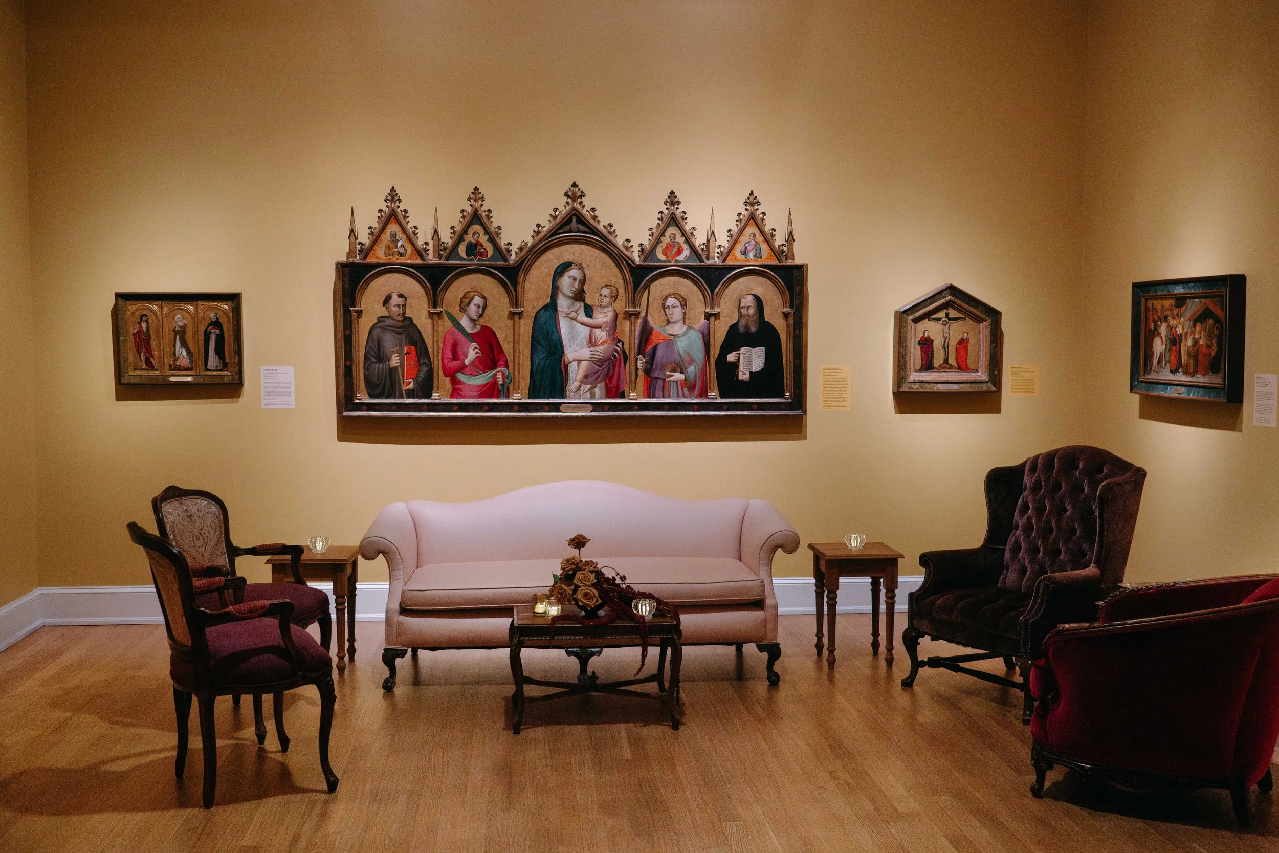 A room with beige walls, hardwood floors, and antique furniture. There is a light pink sofa, two small wooden side tables, and three upholstered armchairs—two burgundy and one black. Several religious paintings, including a central triptych with Chri
