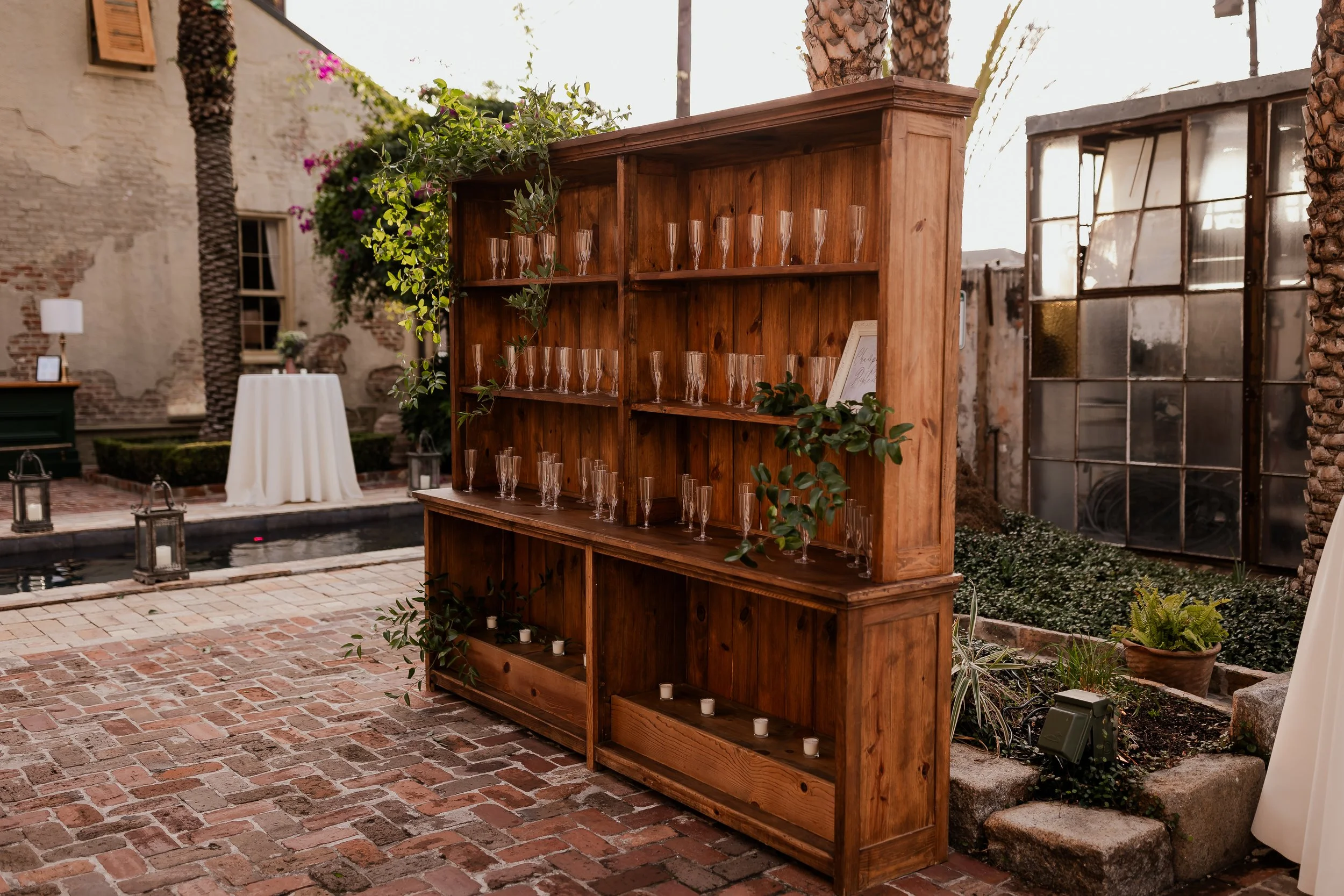 A wooden shelf with glassware, decorated with green foliage, outdoors on a brick patio. There is a white draped table, plants, lanterns, and a distressed brick building in the background.
