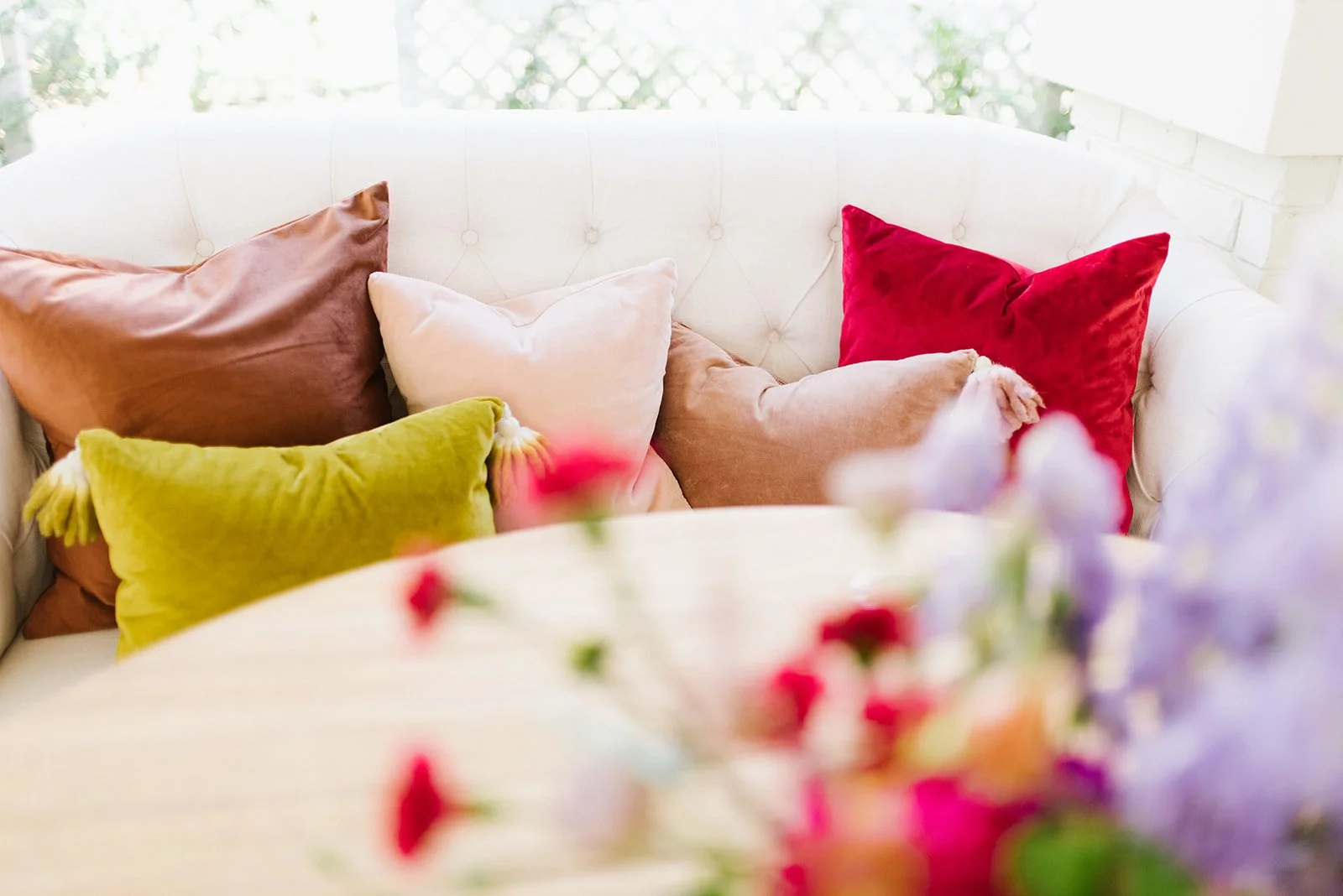 A white tufted sofa decorated with multiple colorful pillows in shades of brown, white, pink, red, and green, with a blurred floral arrangement in the foreground.