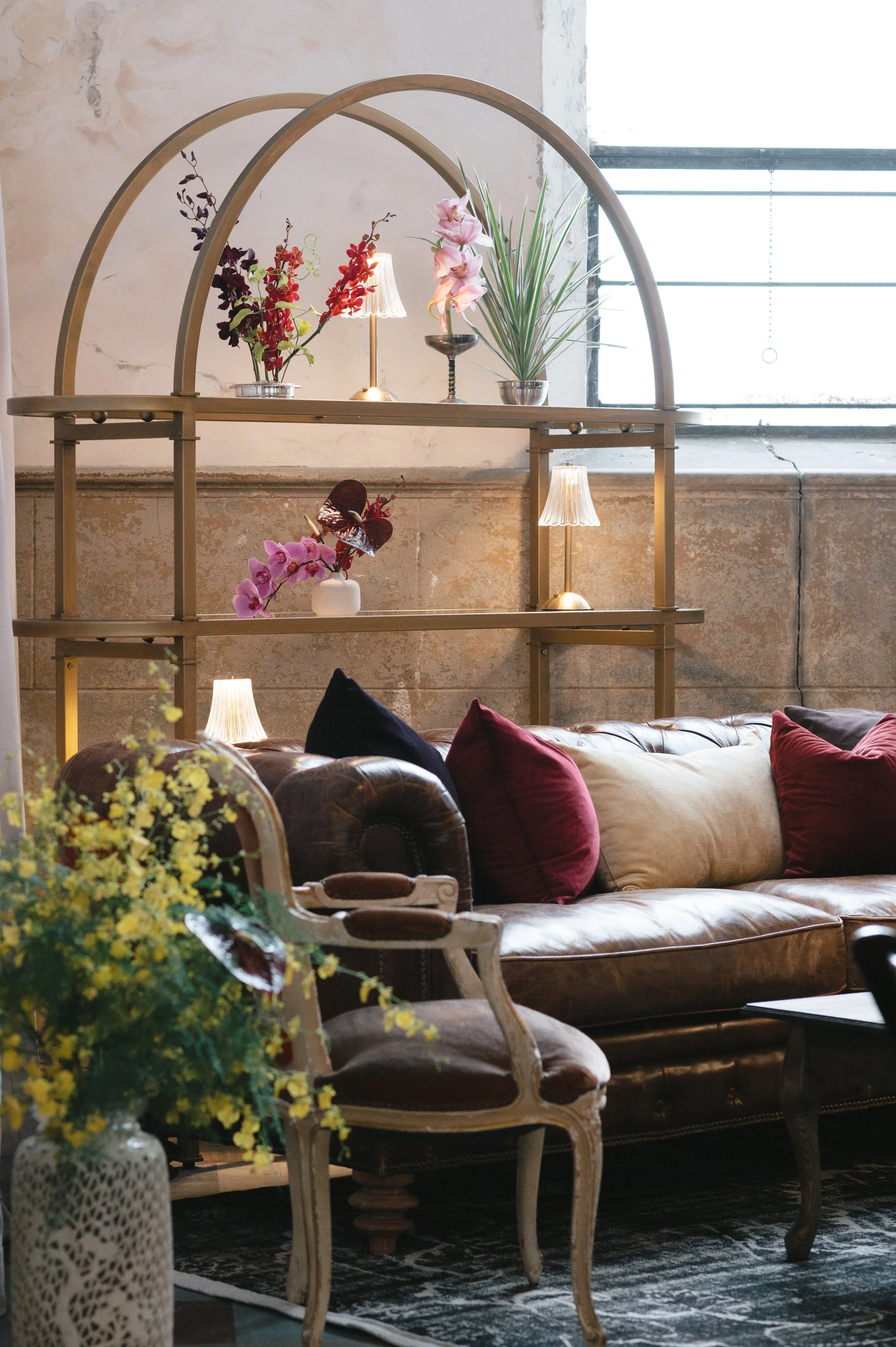 Interior of a cozy living room with a leather sofa, colorful throw pillows, and antique-style chairs. A brass and wood display shelf holds pink and purple orchids and small lamps, set against a rustic wall and large window with natural light.