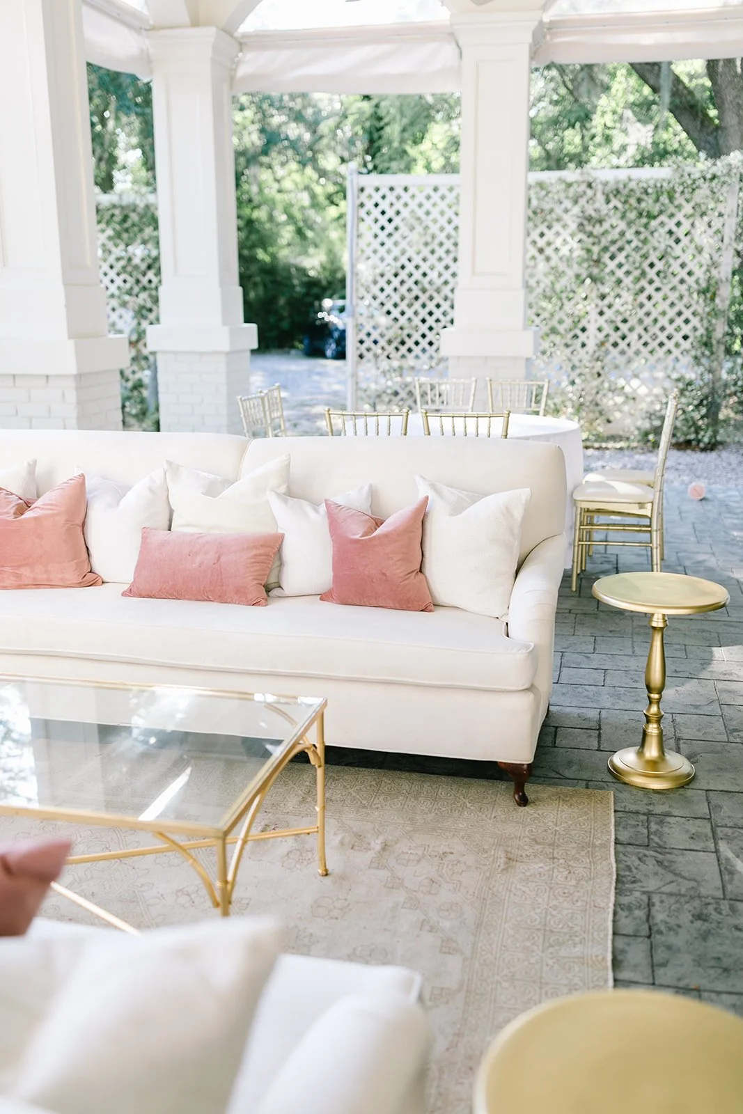 Elegant outdoor patio with white sofa decorated with pink and white pillows, gold side table, glass coffee table, and surrounding chairs and greenery.