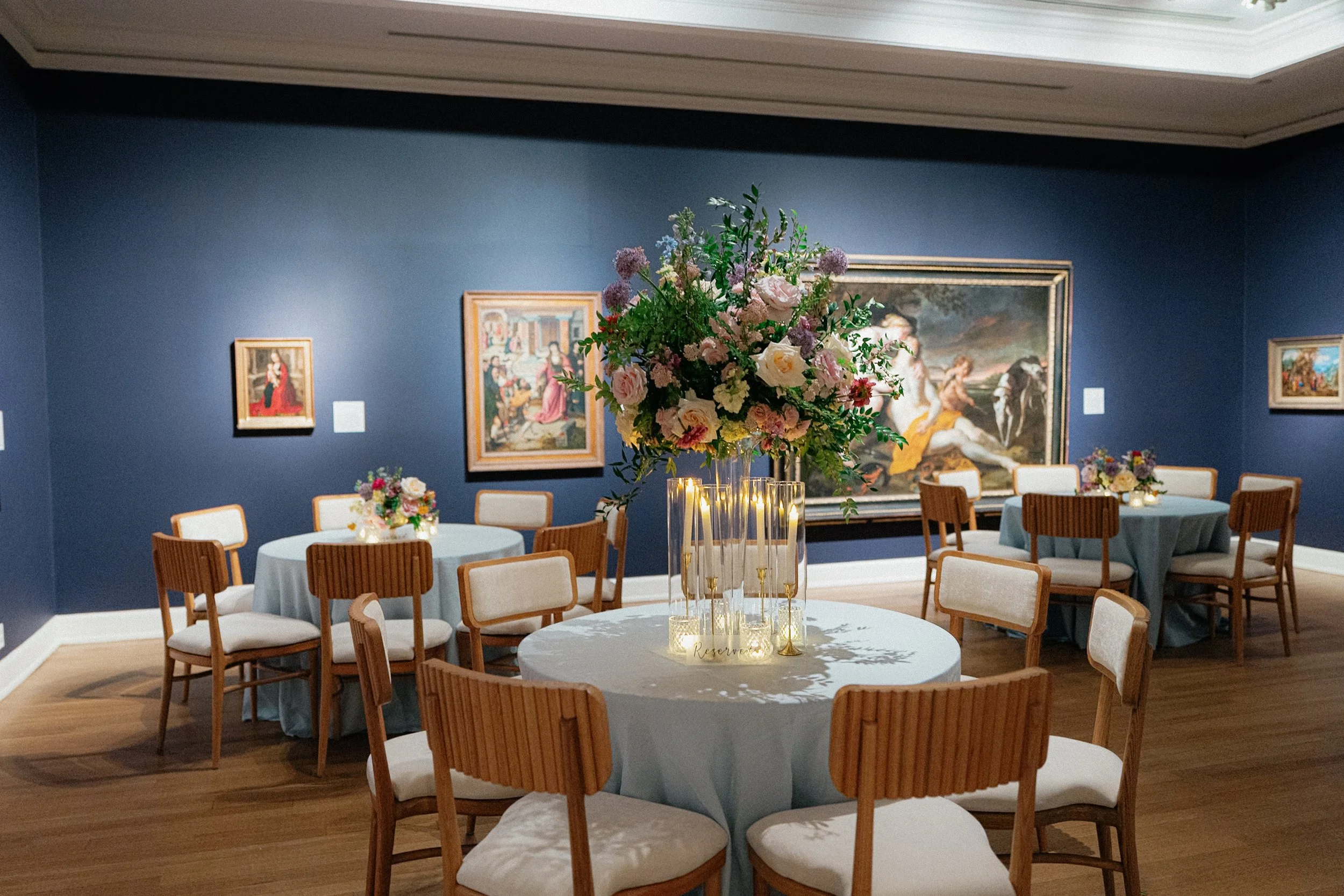 A decorated banquet room with blue walls, several round tables with light blue tablecloths, and wooden chairs with white cushions. The center table has a large floral arrangement with pink and white flowers and green foliage. There are framed paintin