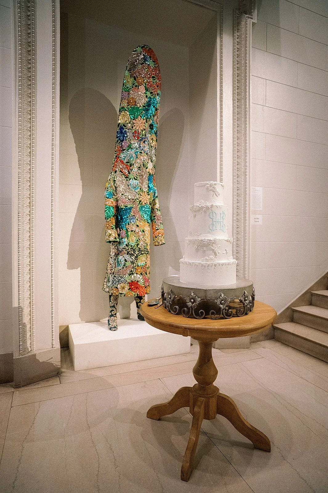 A colorful, floral-embellished dress and a wedding cake on a round wooden table in an art gallery display.