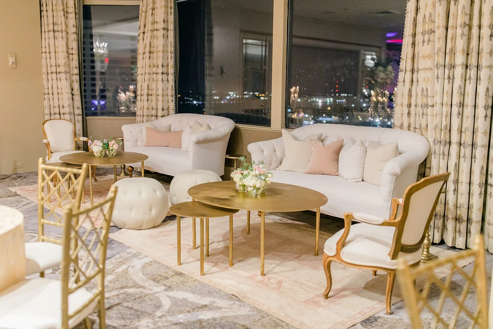 Living room with white sofas, floral pillows, wooden coffee tables with flower arrangements, vintage-style armchairs, twin ottomans, large windows with city skyline at night, patterned curtains, and a decorative rug.