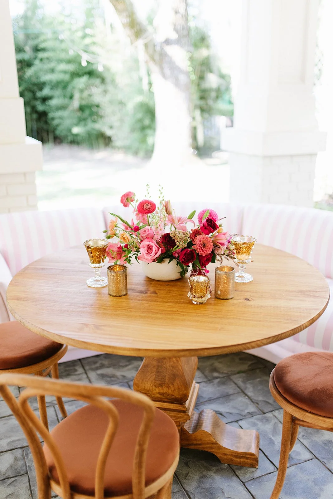 Round wooden table with a floral centerpiece, surrounded by pink and brown chairs, with candles and gold holders, set on a stone floor near a large window with greenery outside.