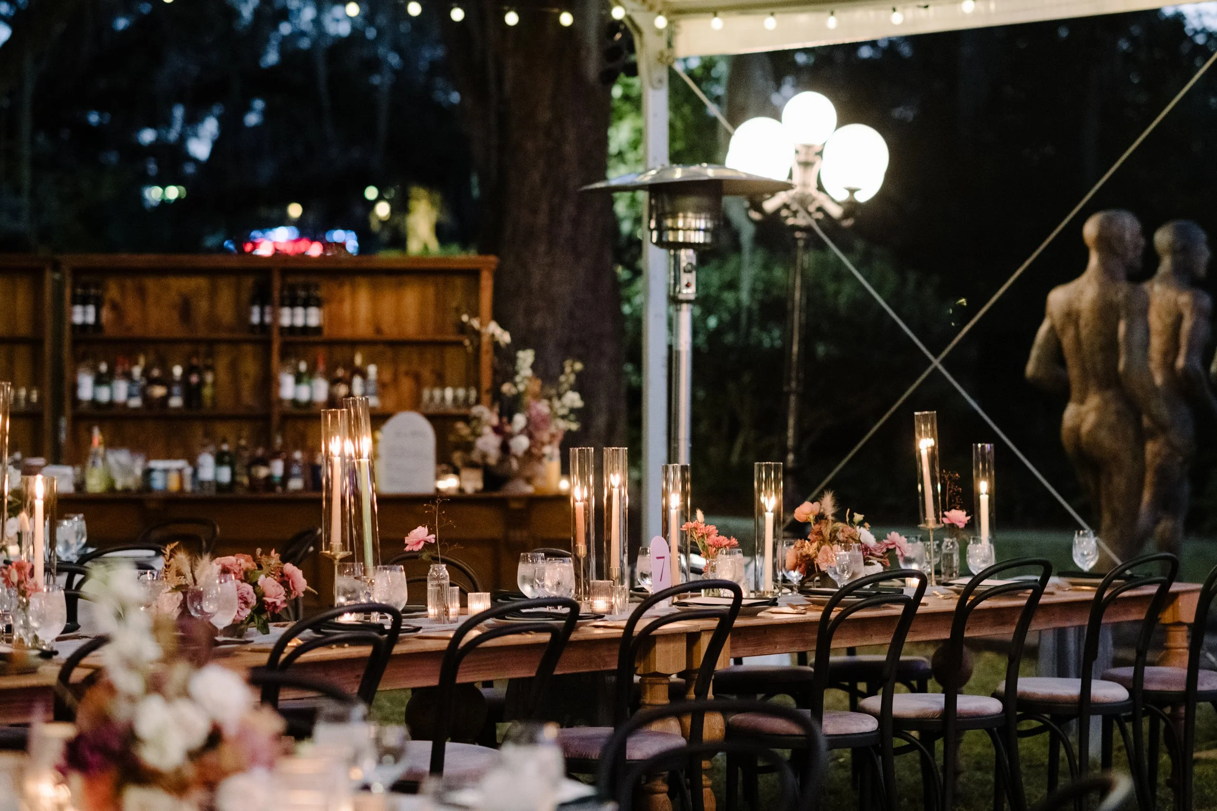 An outdoor event setup at night with a long table decorated with candles and flowers, surrounded by chairs. In the background, a wooden bar shelf with bottles, a large tree, string lights, and decorative bust statues are visible.