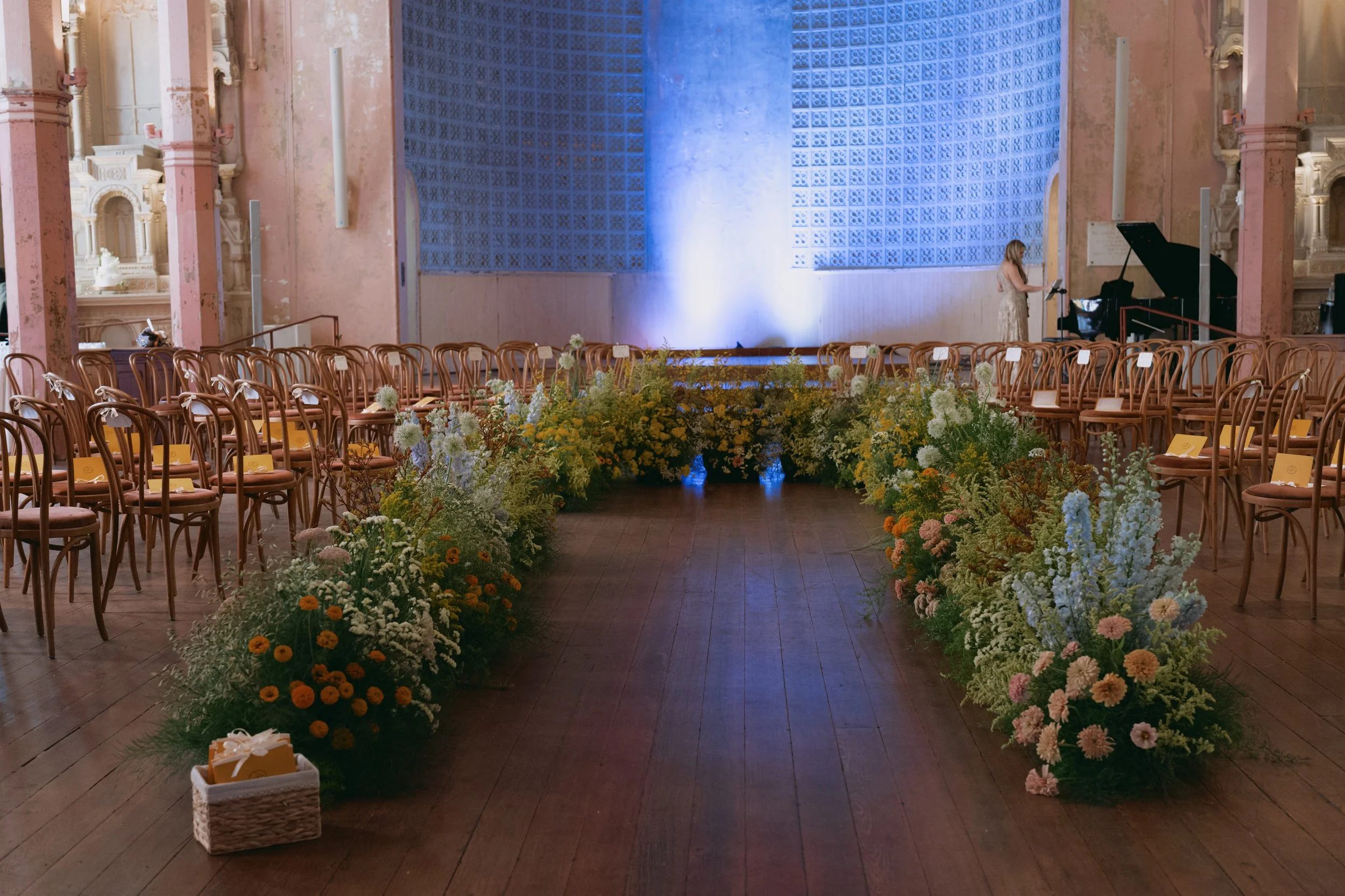 A spacious, elegantly decorated indoor venue with a stage, pink architectural details, and rows of chairs with yellow programs on a floral aisle, preparing for a wedding or special event.