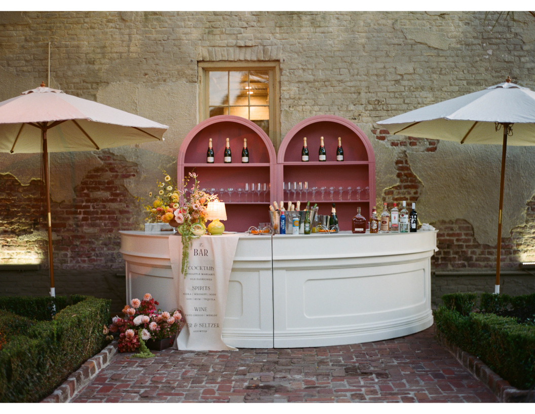Outdoor cocktail bar setup with umbrellas, flowers, and various bottles of alcohol, against an old brick wall with a window.