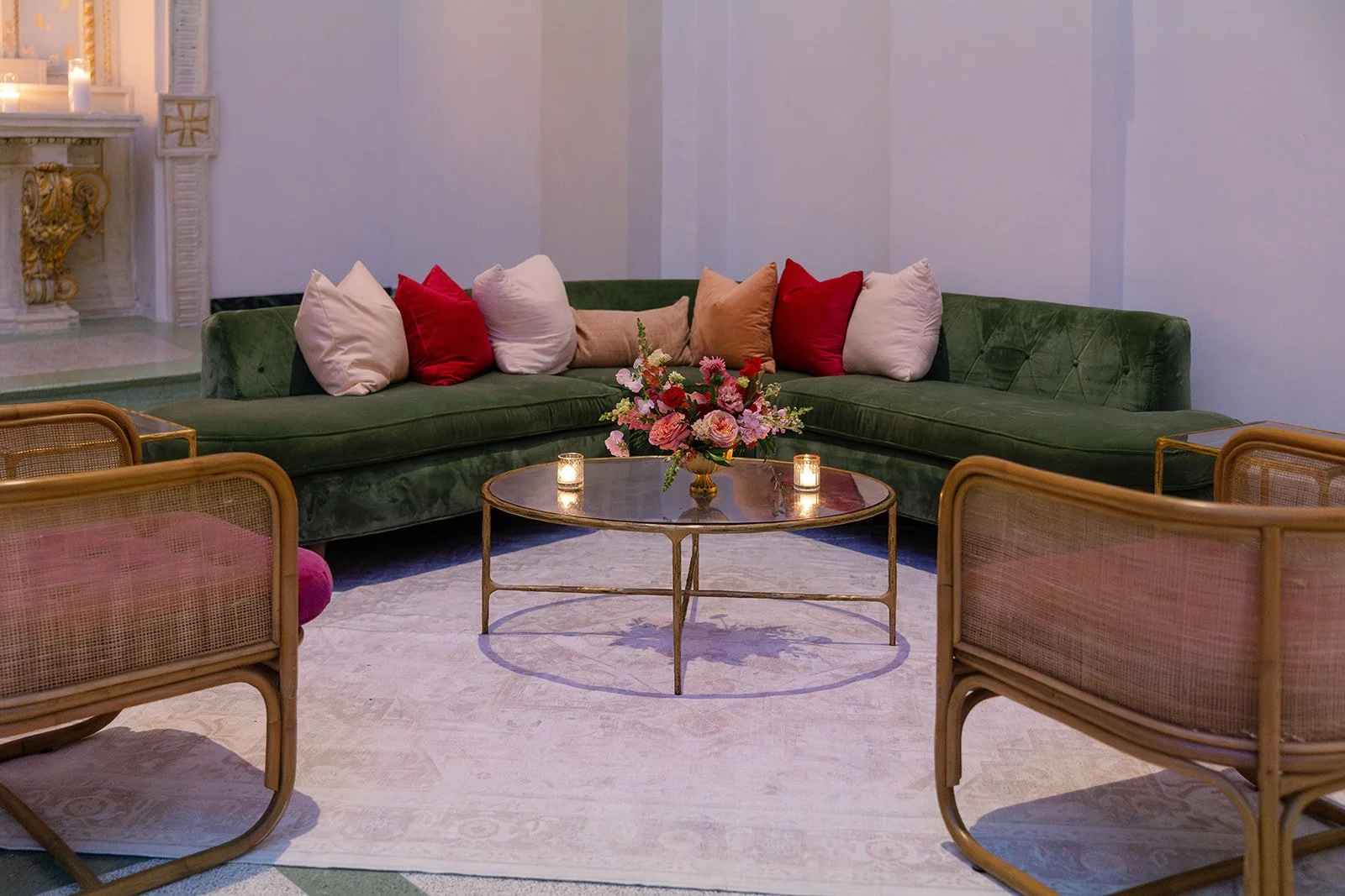 A living room with a green velvet sectional sofa decorated with white, red, and beige pillows. In front, there is a round glass-top coffee table with a floral arrangement, two candles, and wicker chairs surrounding it. Candles are lit, creating a coz