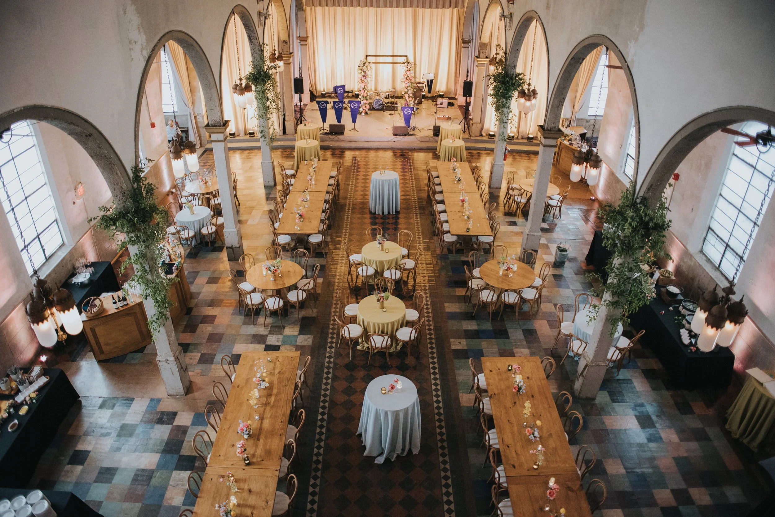 Elegant event space with long tables and round tables decorated with flowers, set up for a celebration or wedding, with a stage at the front draped in light-colored curtains and floral arrangements.