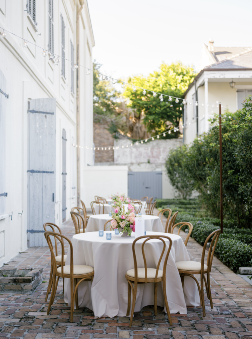 Outdoor dining setup with three round tables covered in white tablecloths, decorated with pink and white floral centerpieces, surrounded by wooden chairs, on a brick patio with string lights overhead, near white buildings and lush greenery.