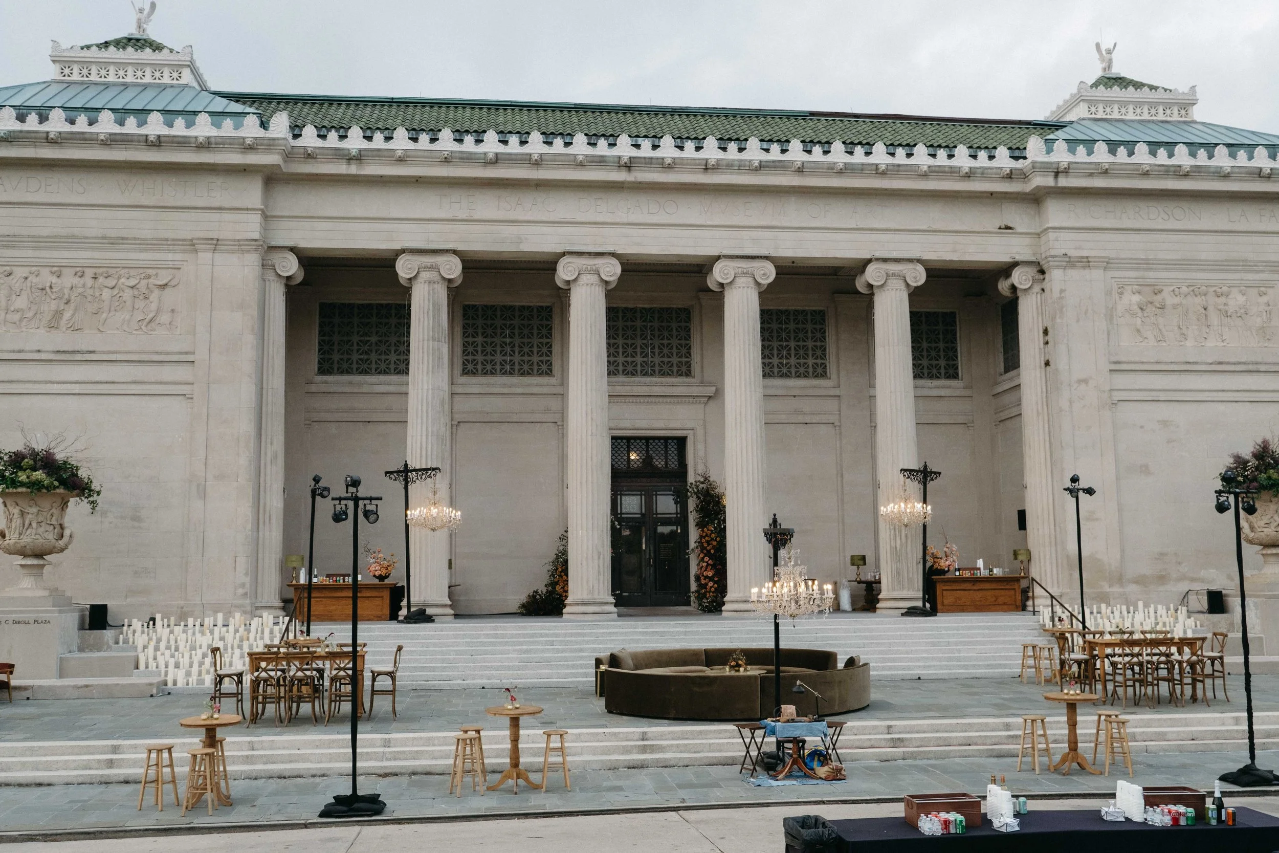 The interior of a grand classical building set up for an event, with tables, chairs, and music stands arranged on steps, chandeliers hanging from the ceiling, and a large statue on the roof.