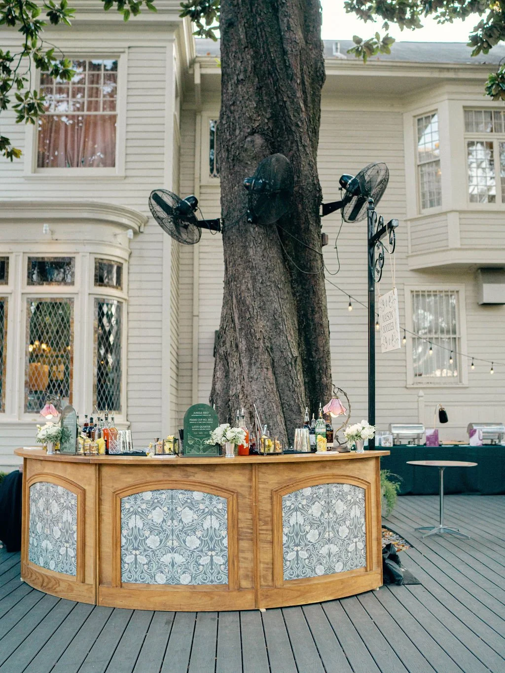Outdoor bar set up on a wooden deck with a large tree in the background, decorated with hanging string lights and a sign. The bar has bottles of alcohol, flowers, and glasses.