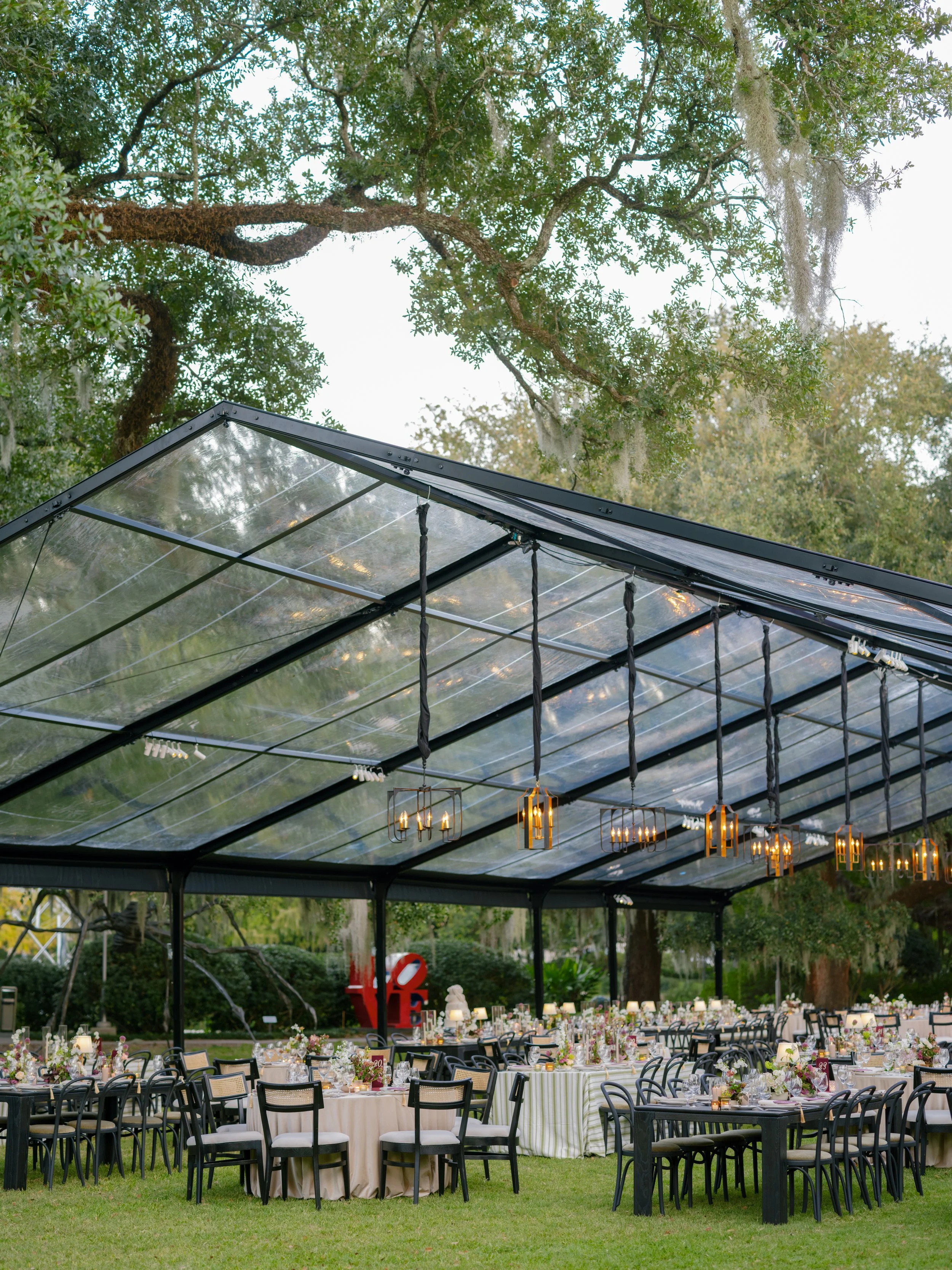 Outdoor event setup with round dining tables covered in floral centerpieces and candles, under a glass pavilion with hanging light fixtures, trees with hanging moss, and a garden background.
