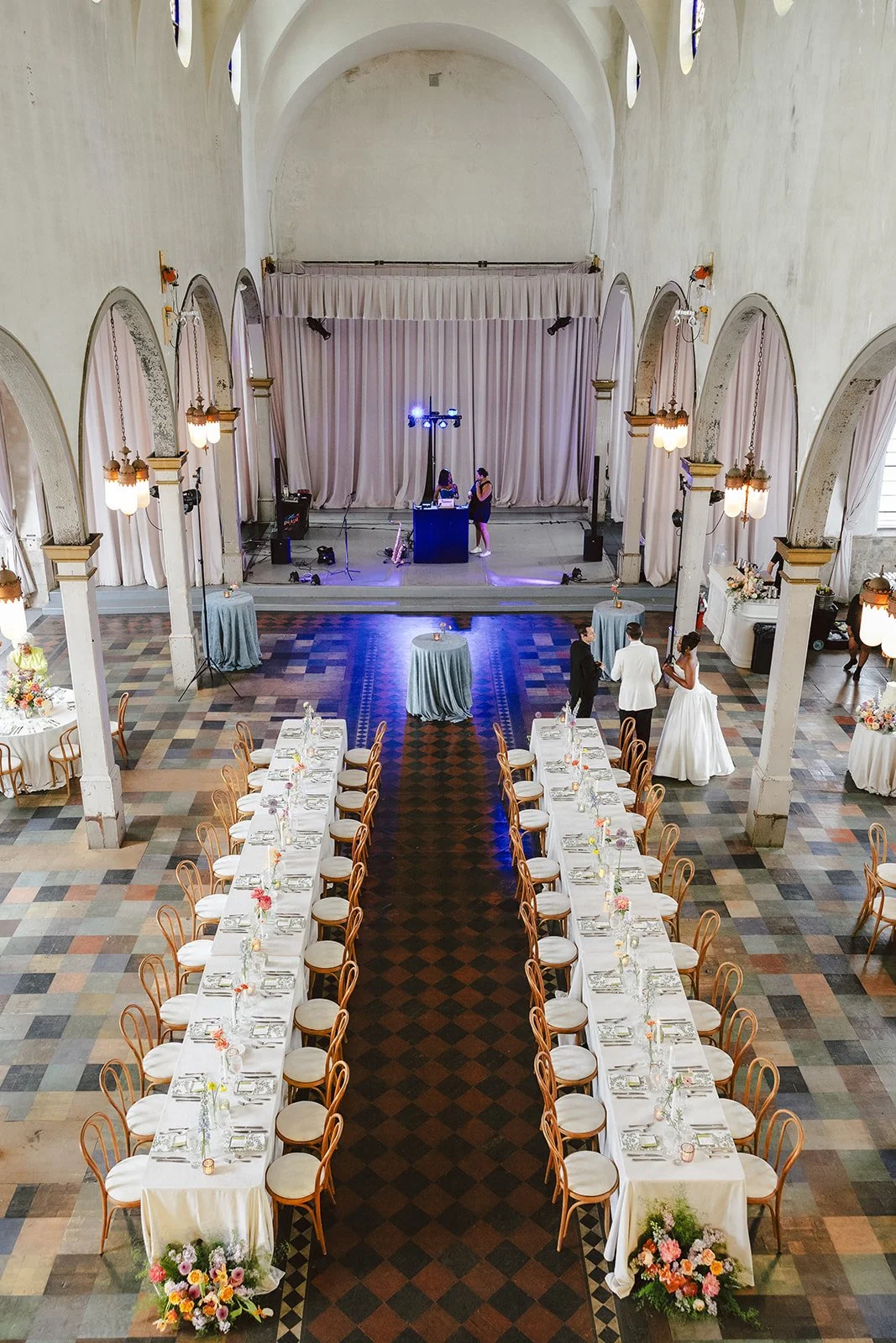 Large event hall decorated for a wedding reception with long tables set for guests, a dance floor in the center, and a stage with a DJ setup at the front. The hall has high arched ceilings, columns, and soft lighting.