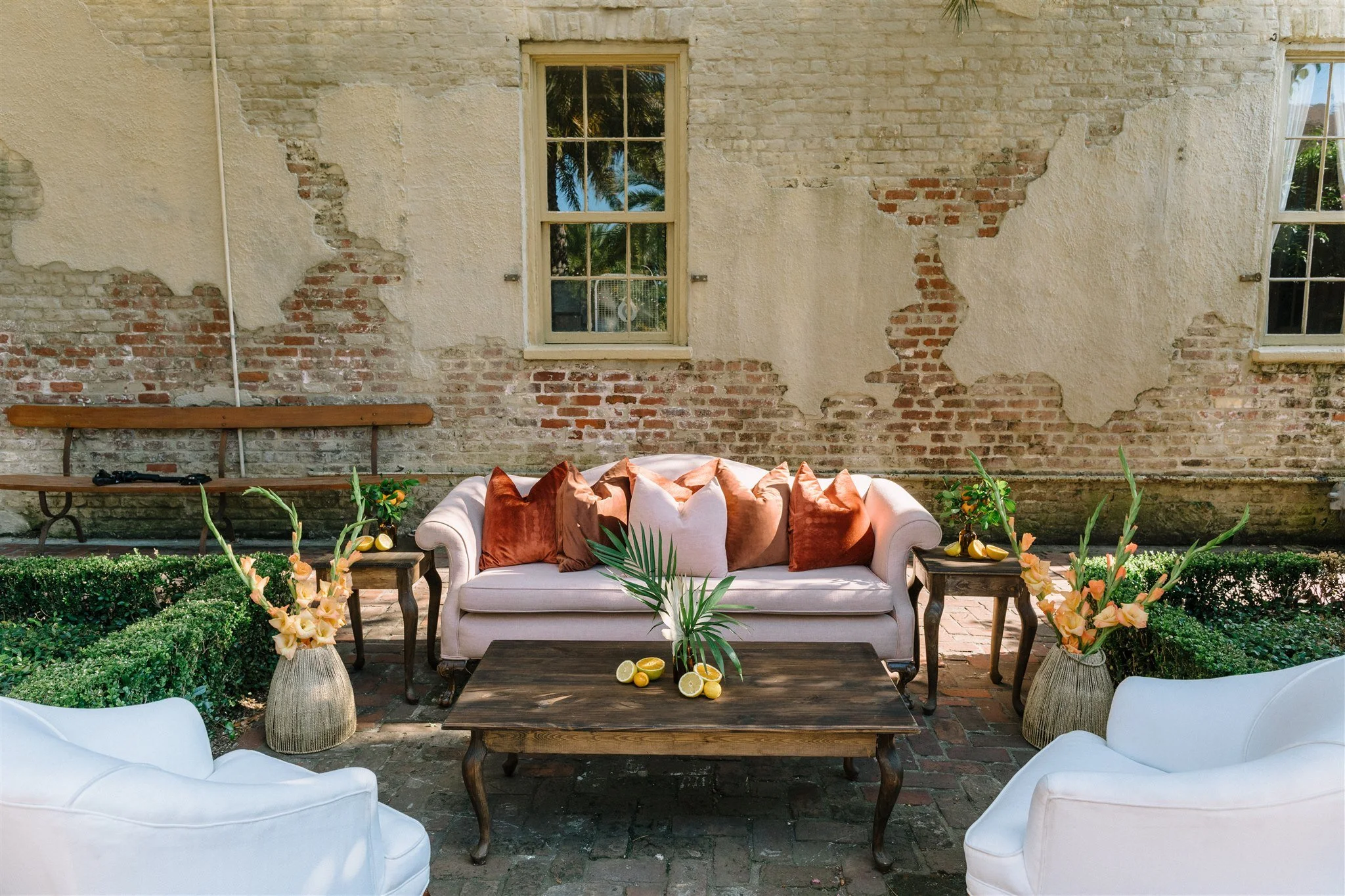 Outdoor patio with white cushioned seating, a wooden coffee table with lemons and greenery, and a vintage brick wall background with three windows, decorated with flowers and plants.