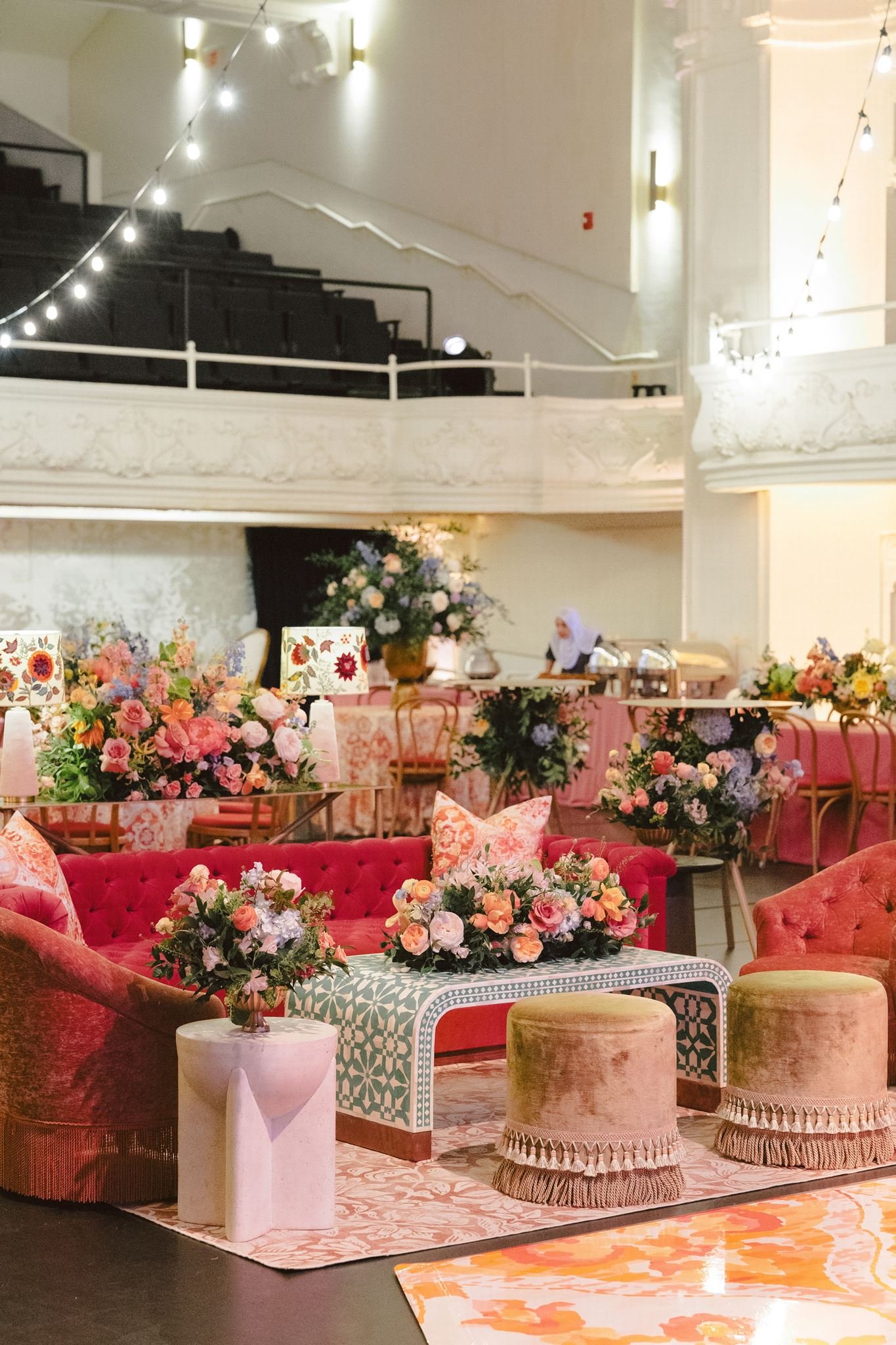 A decorated event space with pink velvet sofas, floral centerpieces, and string lights. There are also tables and chairs, and a person working in the background.
