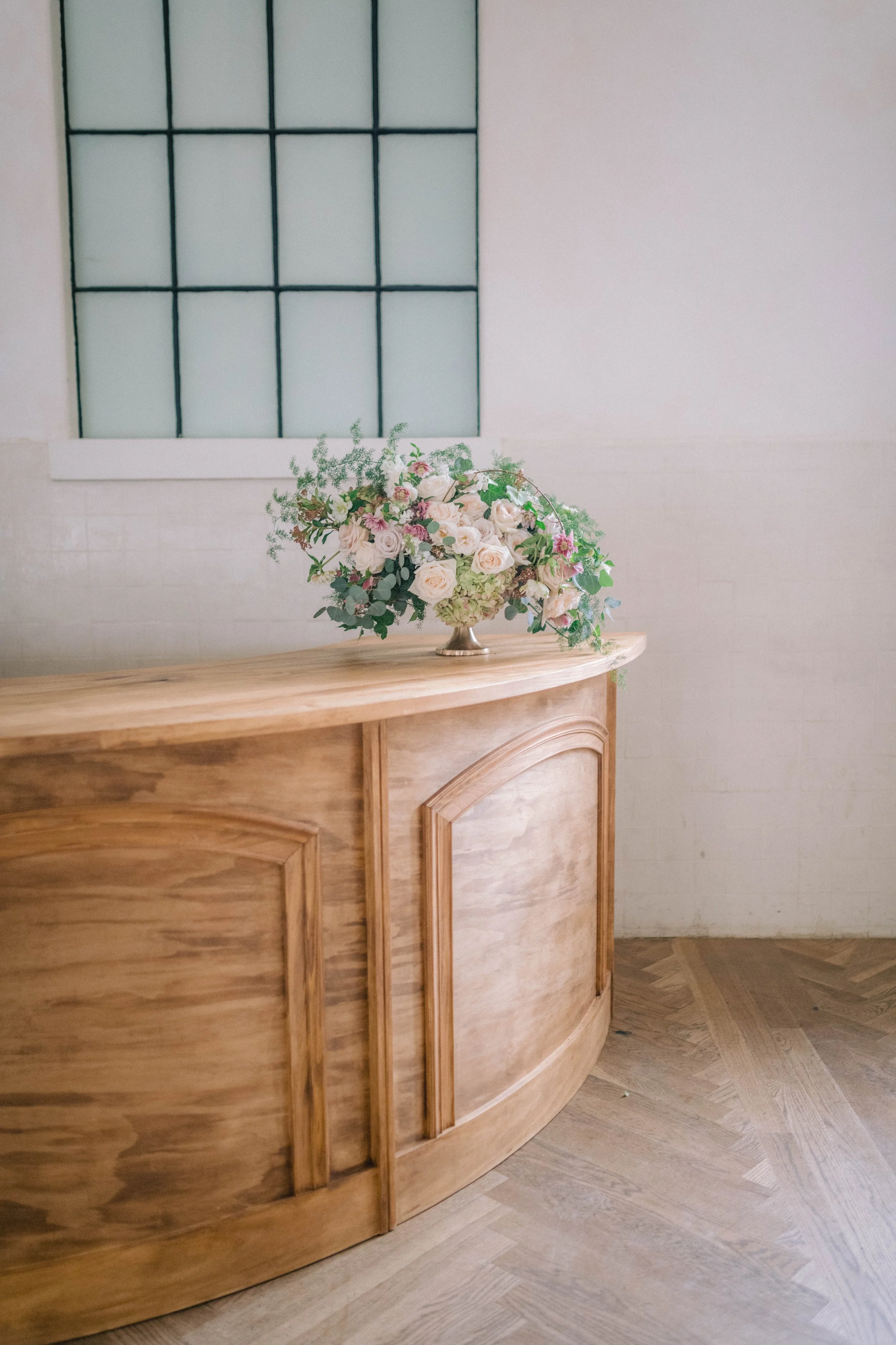 A wooden curved reception desk with a floral arrangement in a silver vase on top, placed in front of a white wall with a large window with black grid-like framing.