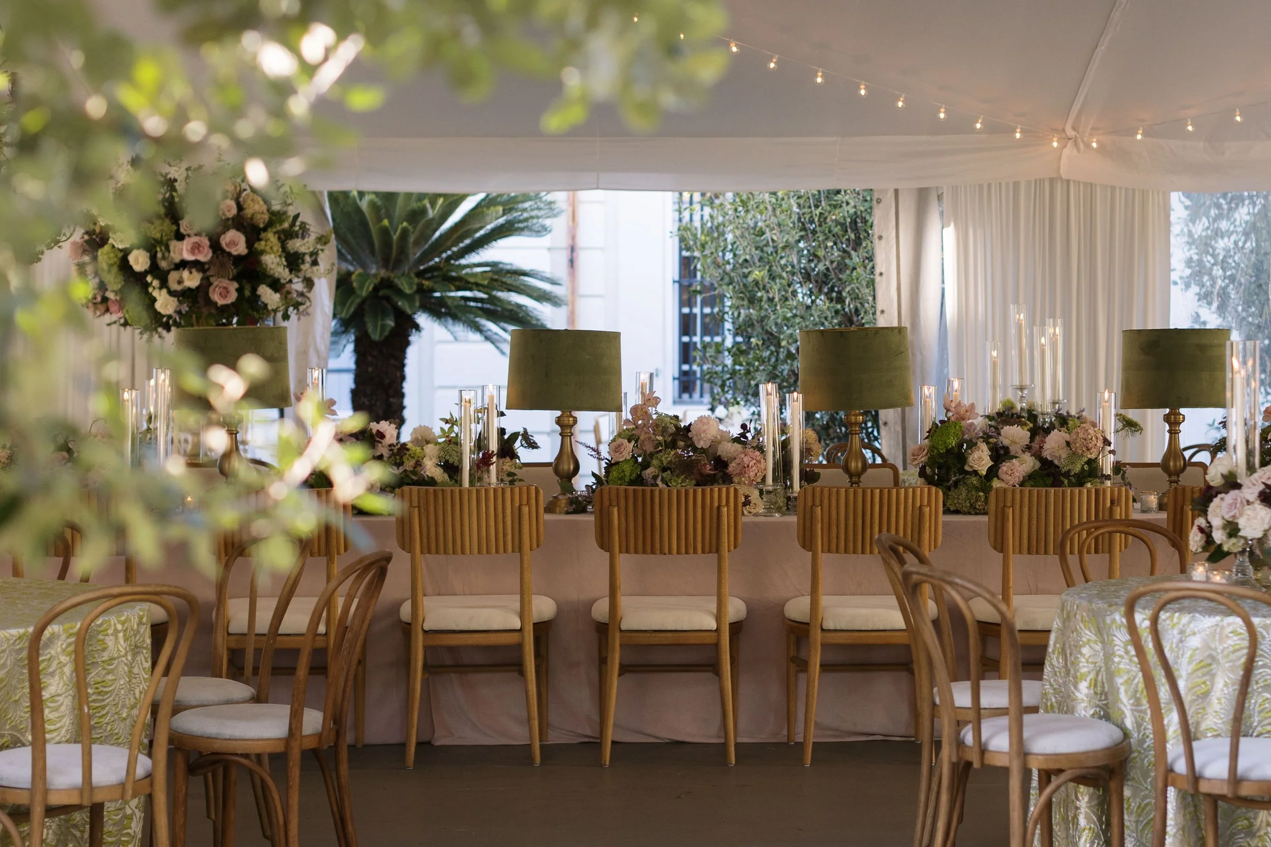 Elegant event space decorated with floral arrangements, candles, and green lamps, with string lights hanging from the ceiling.