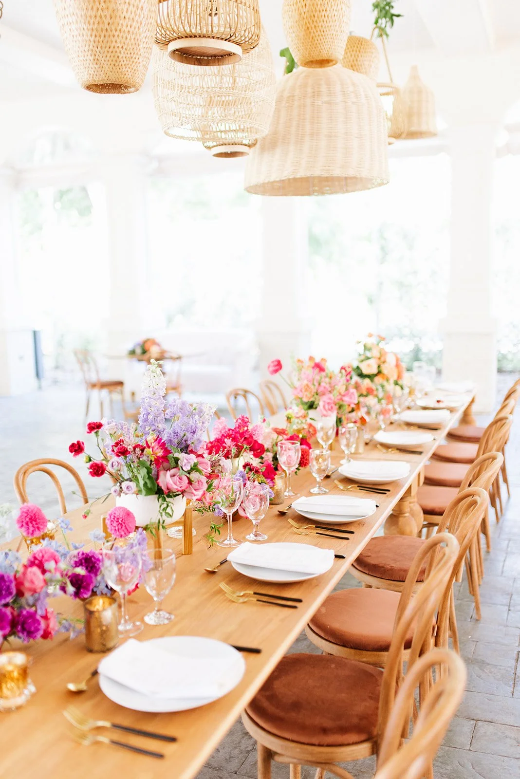 A long wooden dining table decorated with vibrant pink, purple, and peach flowers, set with white plates, gold utensils, and wine glasses in a bright, airy room with hanging wicker light fixtures.