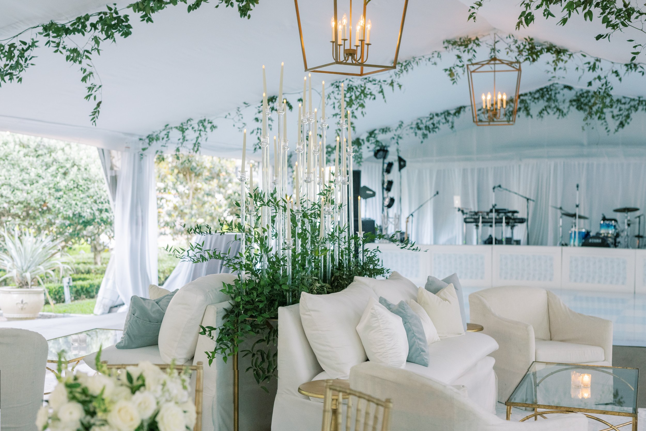Elegant white tent decorated with greenery, hanging lanterns, and tall candlesticks with lit candles for a wedding or event. White couches and gold accent tables inside.