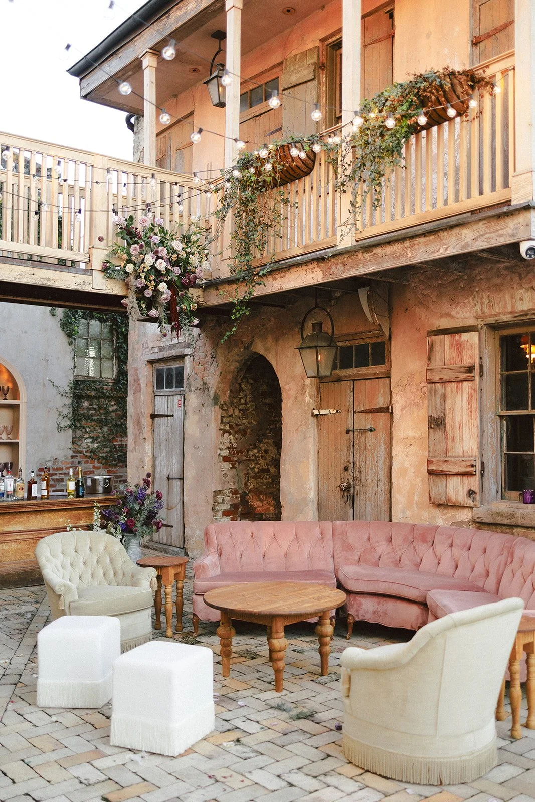 An outdoor patio with vintage furniture, including a pink velvet sofa, white armchairs, and a round wooden table. There's a brick wall with old wooden doors and windows, string lights, and hanging flower baskets on a rustic building. A bar area with 