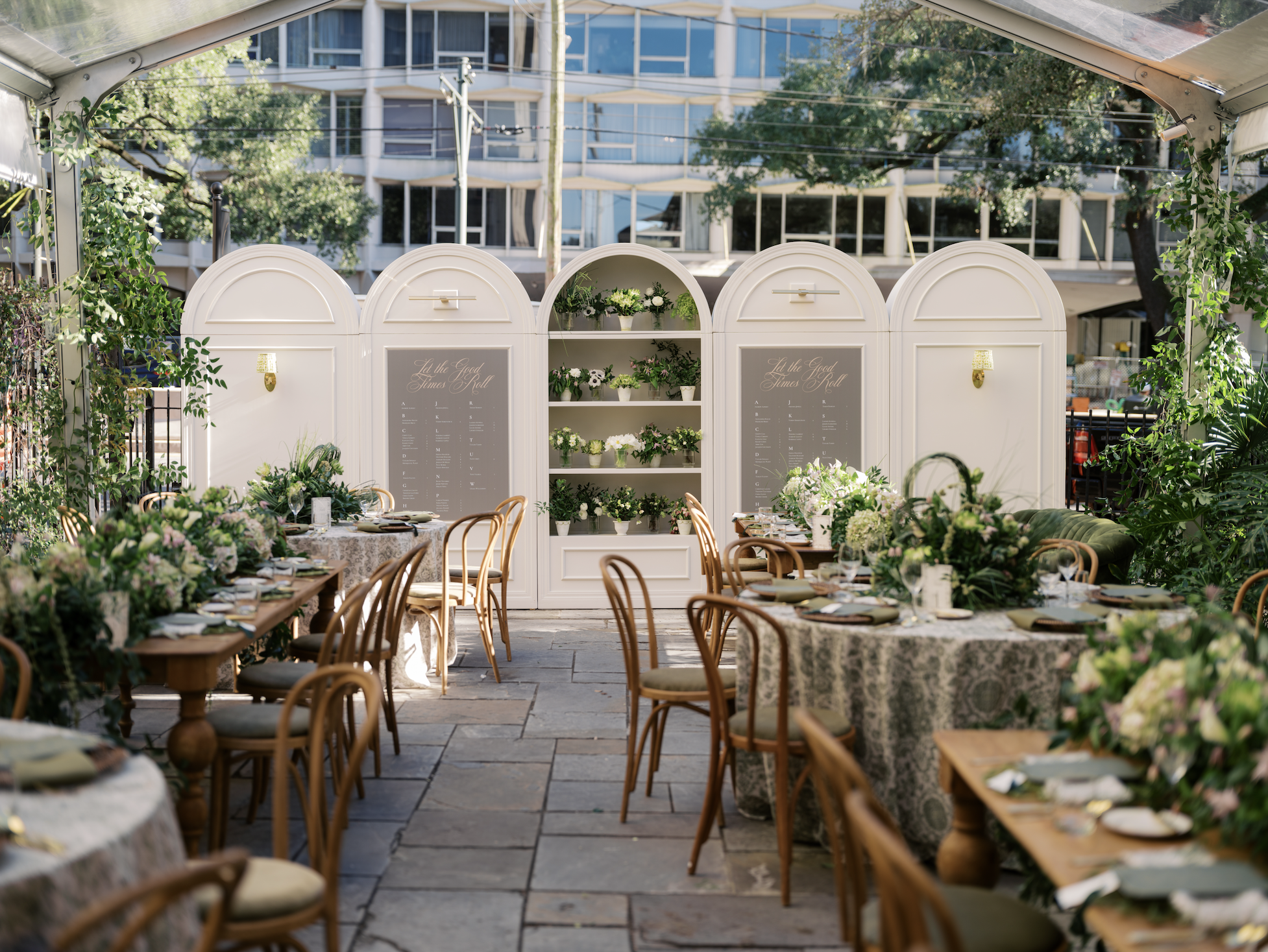 Outdoor event setup with round tables decorated with floral arrangements and green plants, under a transparent tent with string lights, in an urban area with modern glass buildings and trees in the background.