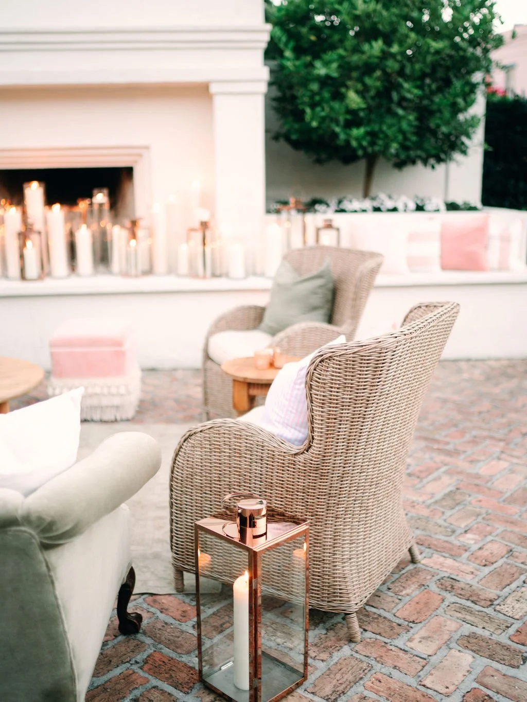A cozy outdoor seating area with wicker chairs, a small side table, candles, and cushions, set against a backdrop of greenery and a fireplace with candles.
