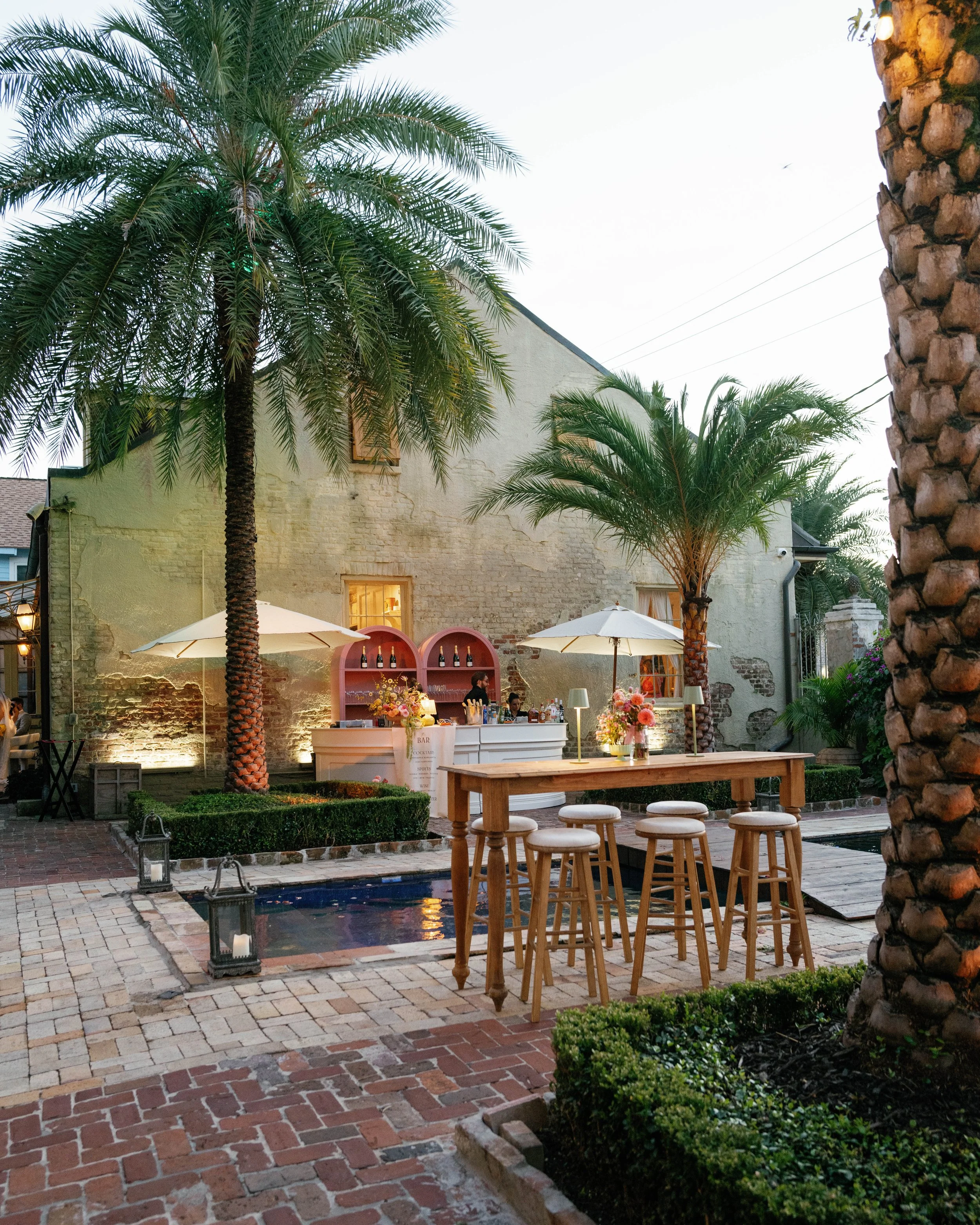 Outdoor patio scene with palm trees, a bar area with bottles and flowers, white umbrellas, and decorative lanterns on a brick and stone paved surface at dusk.
