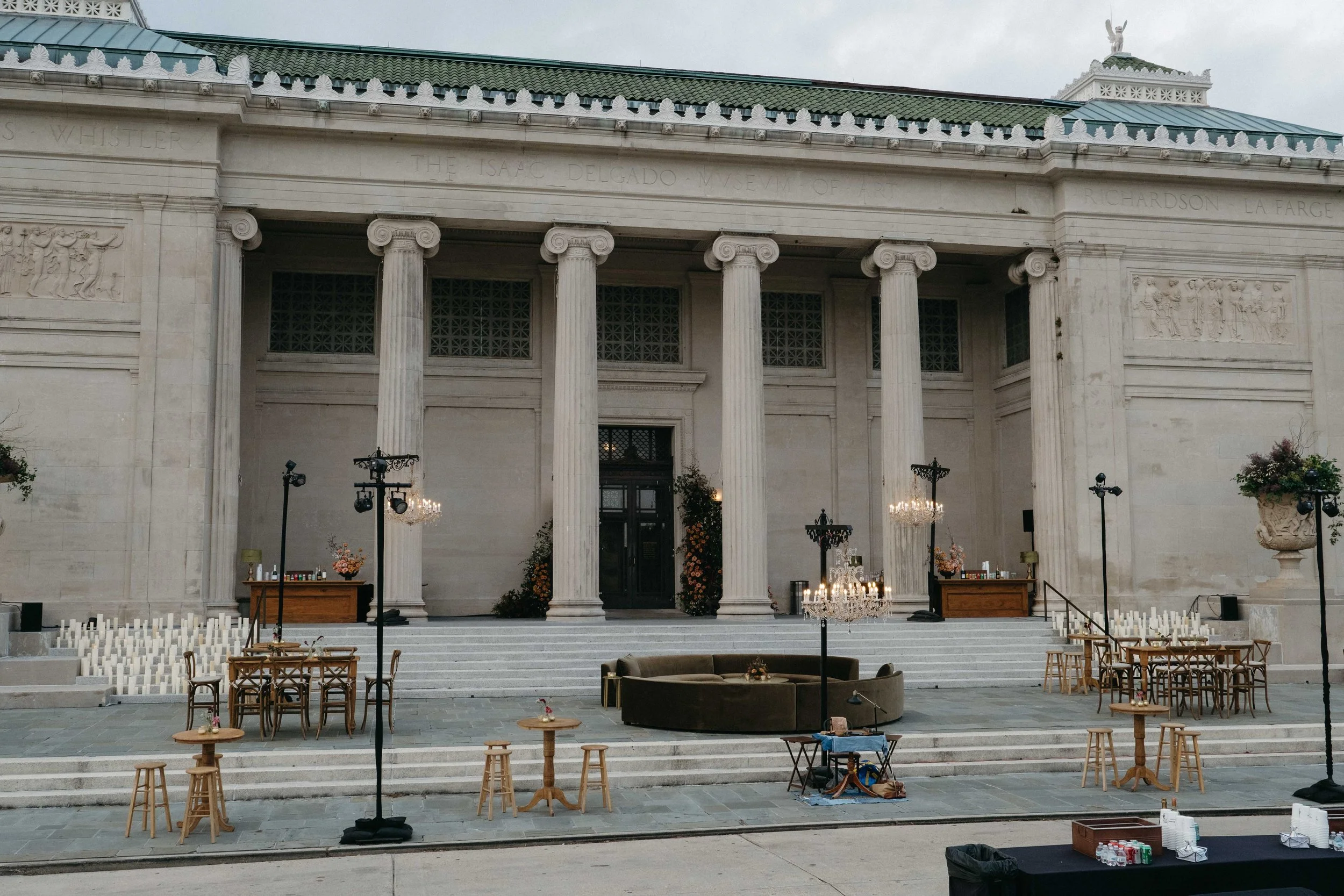 Preparation area set up in front of a classical building with tall columns and engraved frieze, decorated with flowers, candles, tables, and chairs for an event.