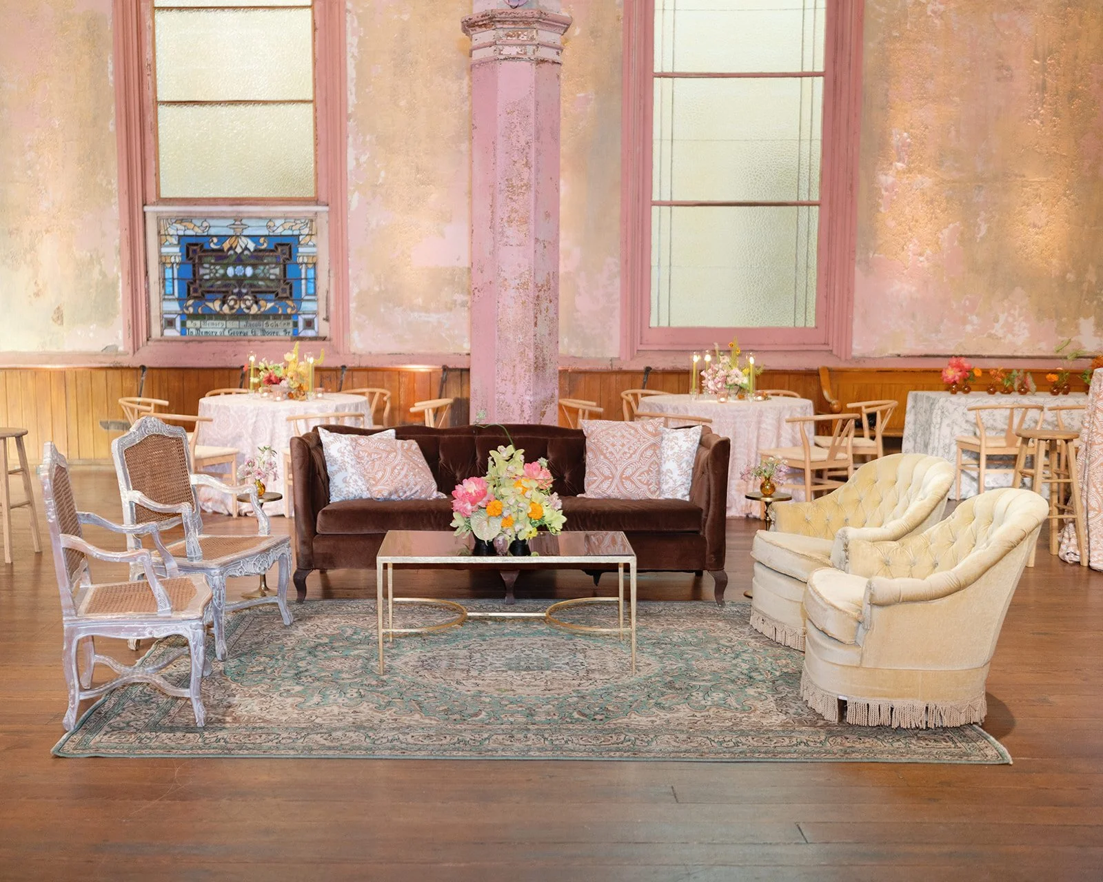 A cozy and elegant living room with vintage furniture, including a brown velvet sofa, two yellow tufted armchairs, and white carved chairs. The room features a floral centerpiece on a glass coffee table, delicate floral arrangements on side tables, a